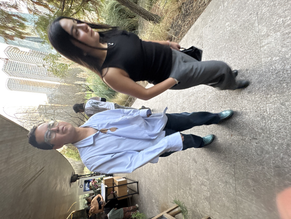 A woman in a black top and gray pants stands near a man with glasses wearing a light blue shirt and dark pants. They appear to be outdoors on a stone walkway, with buildings visible in the background. Other people are present, along with a table and possible signage. The photograph is slightly tilted, creating an unusual perspective.