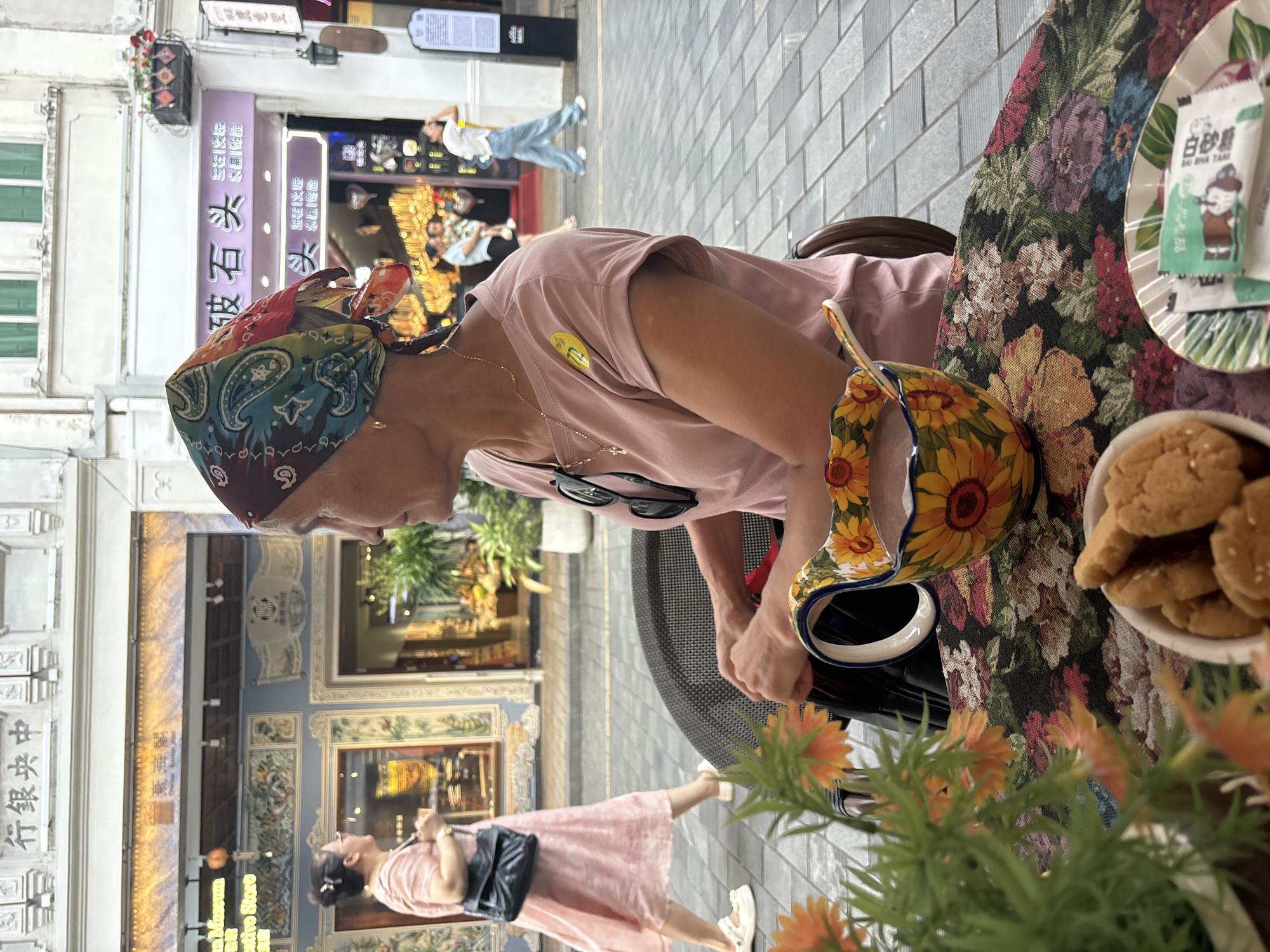 A woman with a colorful bandana on her head sits at a table outdoors in Longhua District, China, facing right and looking away from the camera. The table is covered with a floral tablecloth. In front of her is a large, sunflower-patterned ceramic mug filled with a brown beverage and a spoon. A small bowl of cookies is also on the table, along with a plate containing two packets of sugar. A potted plant with orange flowers is in the foreground. In the background, a street scene unfolds. A woman in a pink dress walks past on the sidewalk, and other pedestrians are visible further down the street, along with storefronts with signage in Chinese characters. The ground is paved with gray tiles. The woman is wearing a pink t-shirt and a gold necklace. Sunglasses are hanging from her t-shirt.