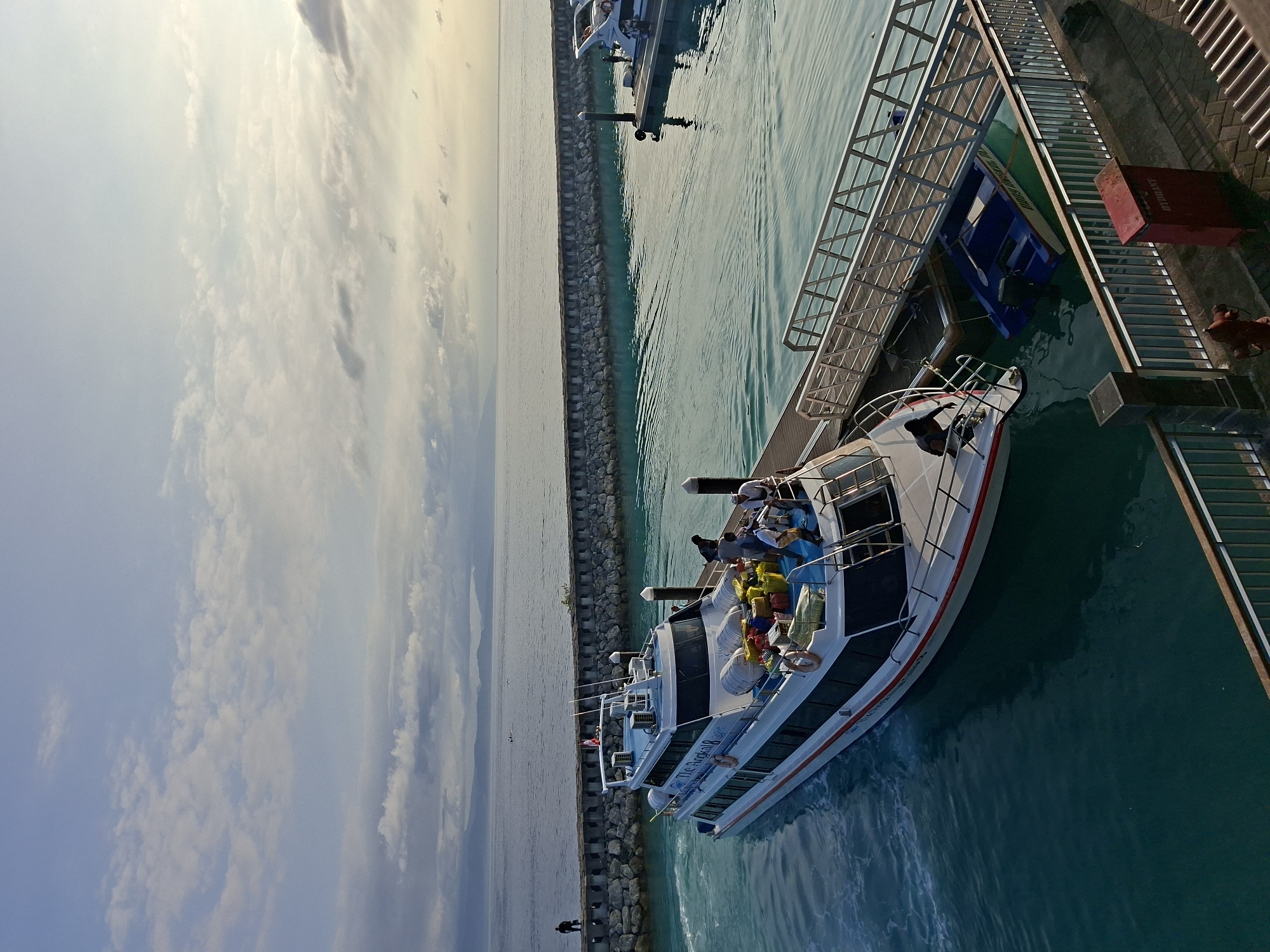The image captures a boat docked at a pier on Nusa Penida, Indonesia. The boat is white with red and blue accents, and passengers are visible on the deck. The pier is constructed of metal and concrete and has a metal staircase leading to the dock. The water is clear and turquoise. The sky is cloudy, with a mix of blue and gray tones. A stone breakwater runs along the edge of the water. A second boat is docked further out on the water.
