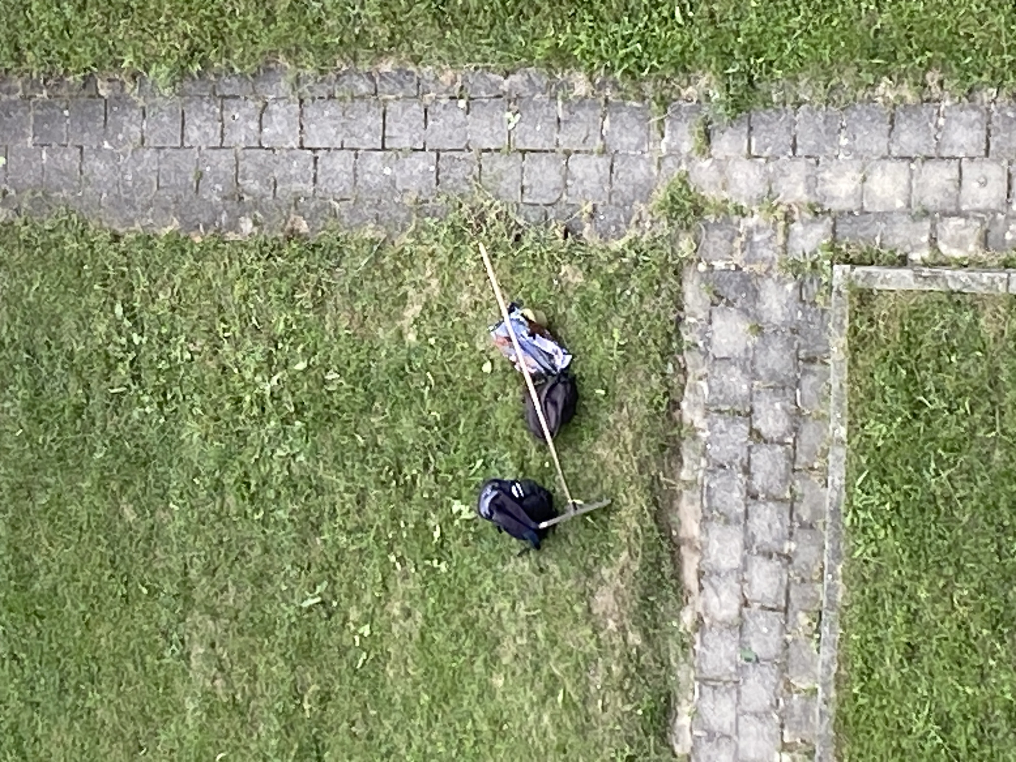 Two black backpacks are lying on a patch of grass next to a stone pathway. One backpack, to the left, is upright and appears to be a standard hiking backpack with straps visible. The other backpack, to the right, is lying on its side and is partially covered by what appears to be a blue and white patterned umbrella or bag. A thin, light-colored stick or pole extends horizontally across the image, connecting the two backpacks, with its ends resting near each bag. The pathway is composed of rectangular gray stone pavers arranged in a grid pattern and is bordered by green grass. The scene is observed from a high vantage point, looking down.