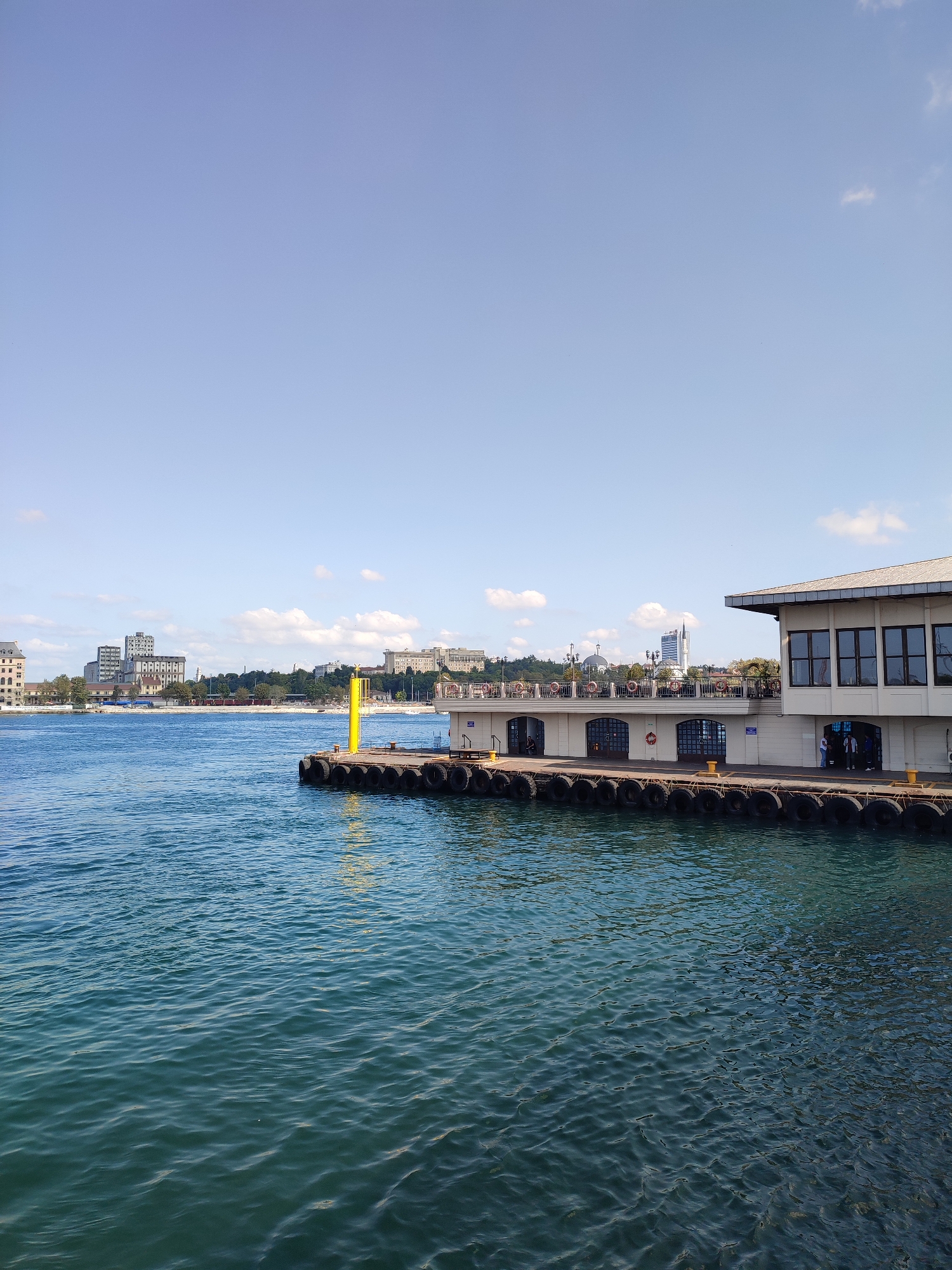 The image depicts a waterfront scene in Kadıköy, Turkey, with a clear blue sky and calm turquoise water. In the foreground, the water exhibits gentle ripples. A ferry terminal building with a white facade and large windows stands to the right of the frame, projecting into the water. The terminal's exterior is lined with a row of tires serving as a protective buffer against boats. Adjacent to the terminal, a bright yellow bollard is visible. Across the water, the Kadıköy skyline is visible, featuring various buildings, including residential blocks and a prominent white dome structure, with a cluster of trees and greenery on the embankment. Several people are casually present at the ferry terminal entrance.
