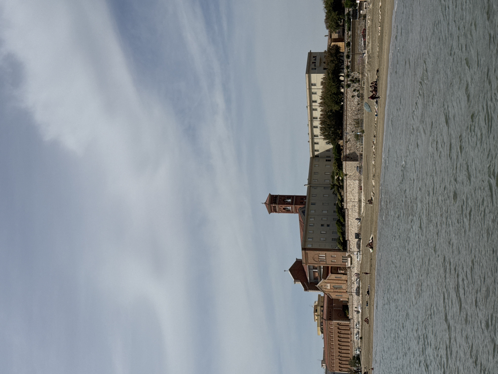 The image shows a coastal scene in Nettuno, Italy. The sky is filled with streaks of white clouds against a light blue background. Buildings line the beach, featuring varied architectural styles and colors, mostly in shades of brown and red brick, with some lighter colored structures. A tall brick tower is visible among the buildings. People are sparsely scattered along the sandy beach, with some appearing to be resting or interacting. A body of water, likely the Tyrrhenian Sea, fills the right side of the image. The buildings are separated from the beach by a narrow strip of greenery and a low stone wall.