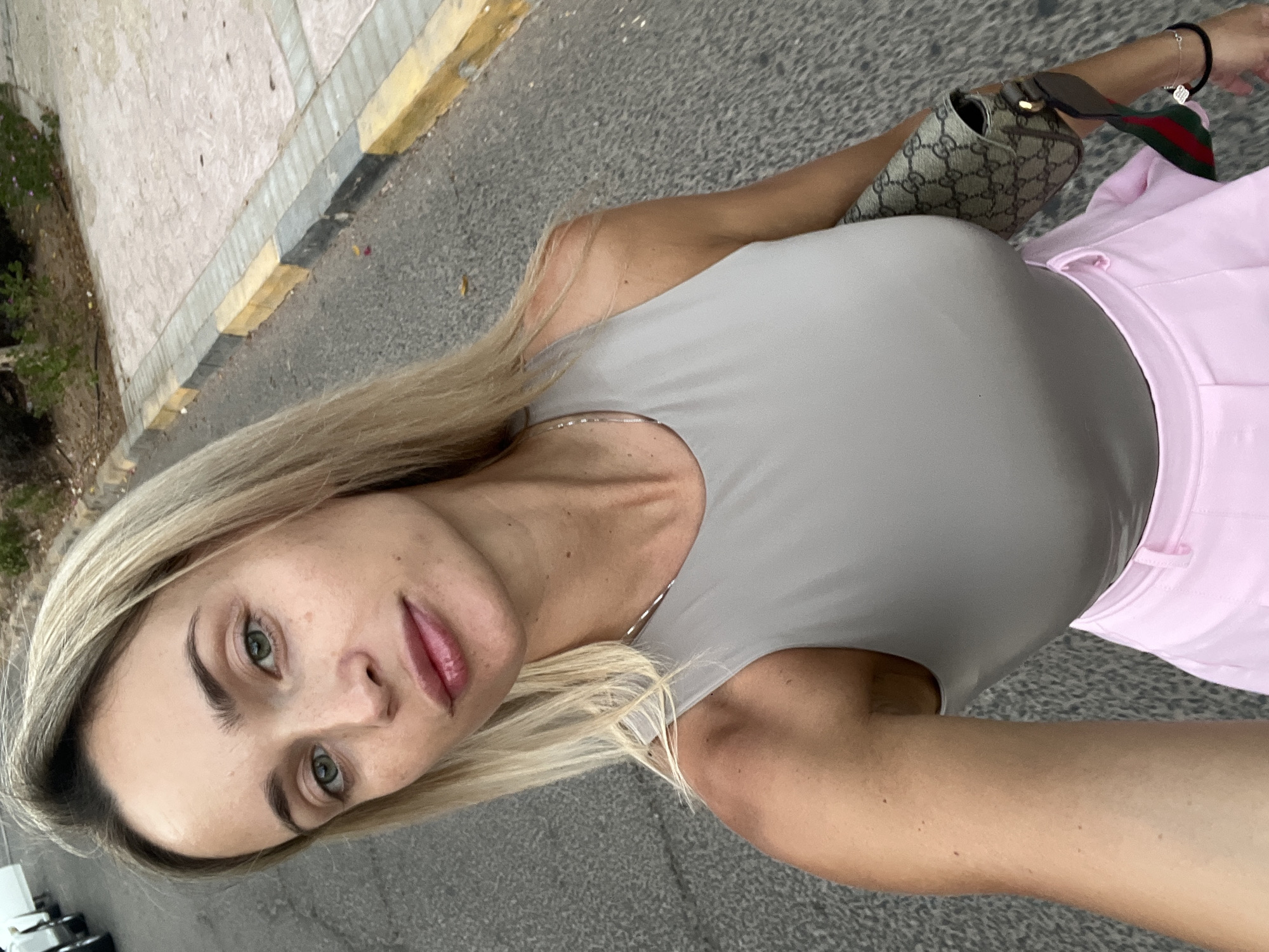 A blonde woman stands on a paved street, taking a selfie. She is wearing a grey tank top and a pink skirt. A patterned handbag with a red and green strap is slung over her shoulder. The street is lined with a concrete curb, partially painted yellow and grey. In the background, there is dry brush and some small plants. The scene appears to be in Sharm El Sheikh, Egypt.