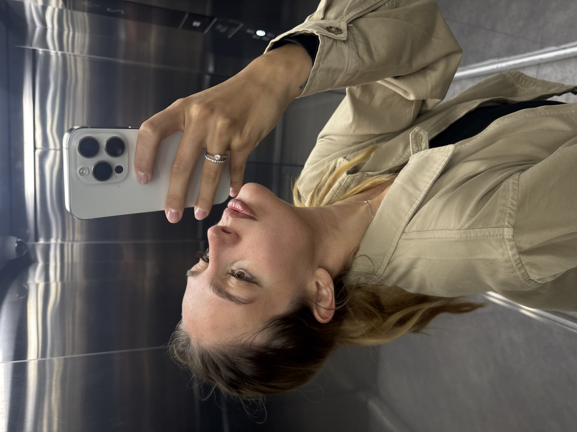 A woman in Unknown, Unknown, takes a selfie in an elevator. She holds a white iPhone in her left hand with a silver ring on her ring finger. She is wearing a beige jacket over a black top. The elevator has stainless steel walls.