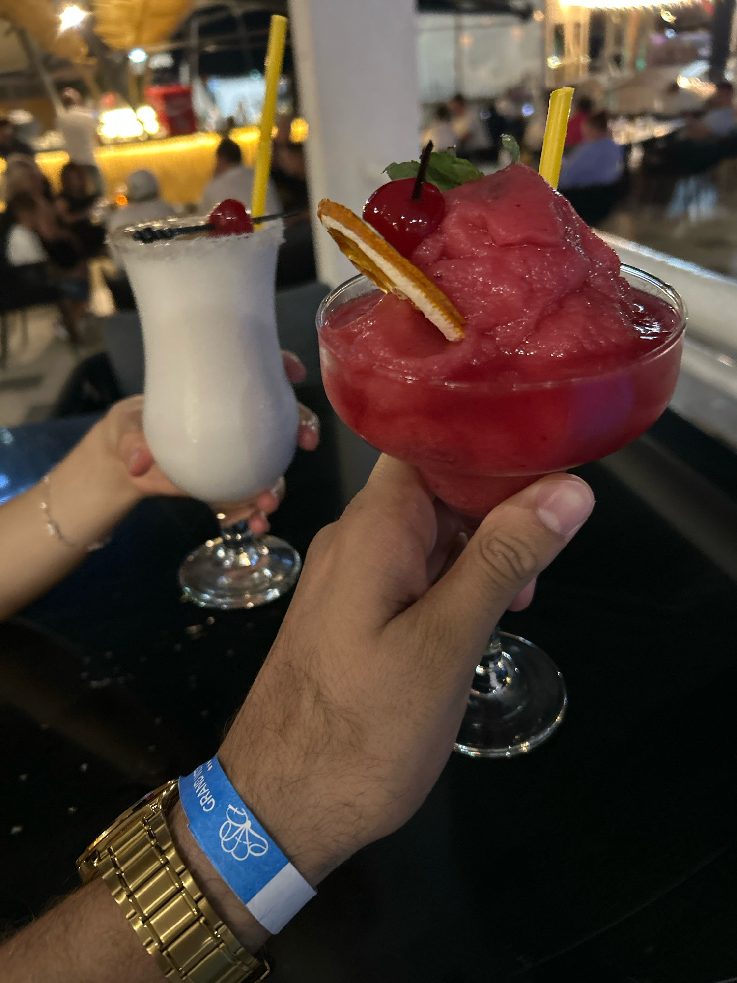 Two people are holding cocktails at a bar in Nesebar, Bulgaria. The person in the foreground, on the right, is holding a frozen pink cocktail in a margarita glass, garnished with a cherry, mint, and a slice of orange. They are wearing a gold watch and a blue wristband. The person in the background, on the left, is holding a white cocktail in a hurricane glass, garnished with a cherry and a yellow straw. The bar appears to be outdoors and busy, with other patrons visible in the background. The lighting is dim and atmospheric, with yellow and white lights illuminating the scene.