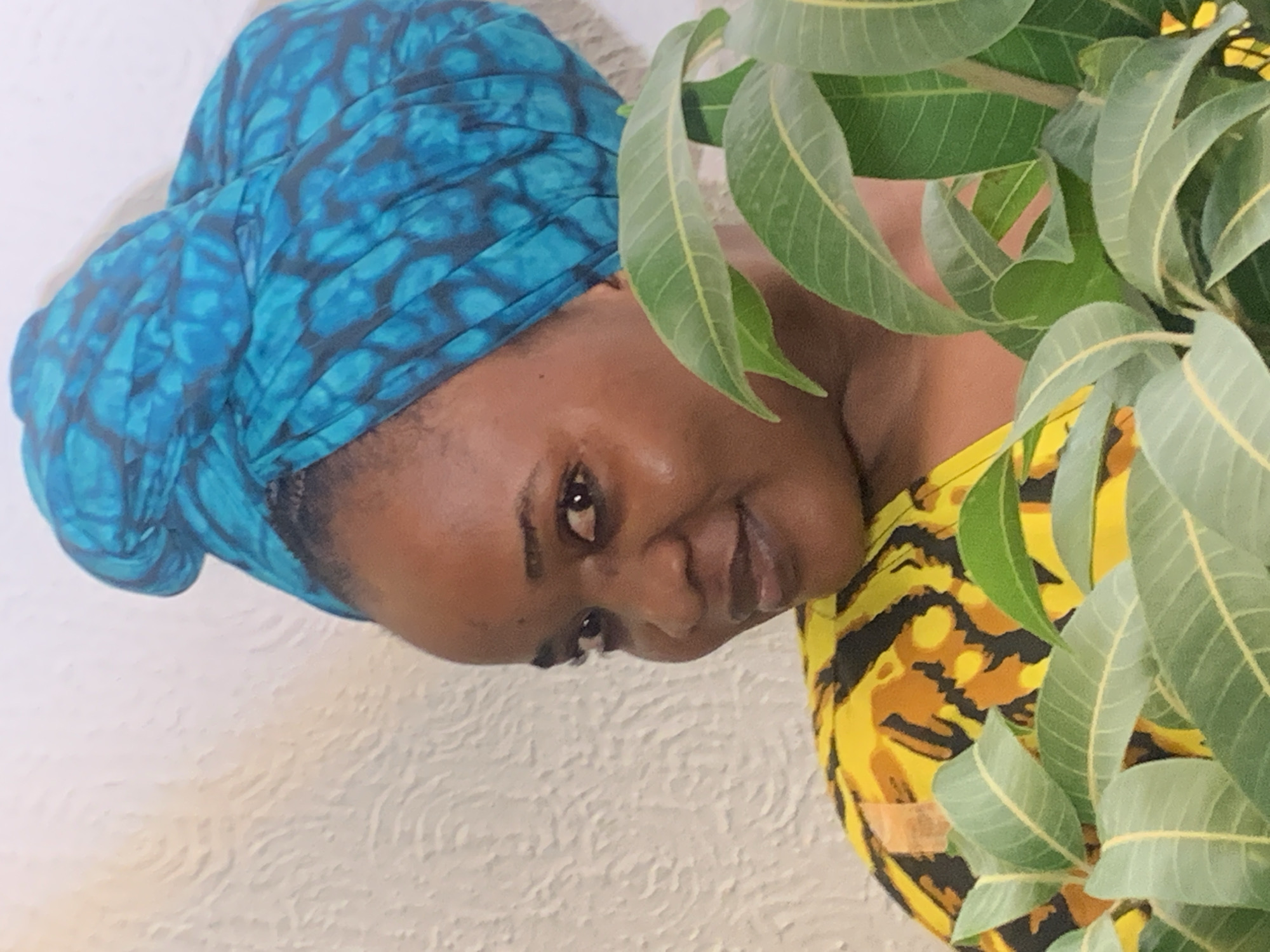 A woman with brown skin is facing the camera at an angle from the upper left corner of the image. She is wearing a blue and black patterned headwrap, and a yellow and black patterned shirt. Part of the background behind her is a white textured wall. Leaves are also present on the right side of the frame. The location is Barakin Akawo, Nigeria, as per the prompt.