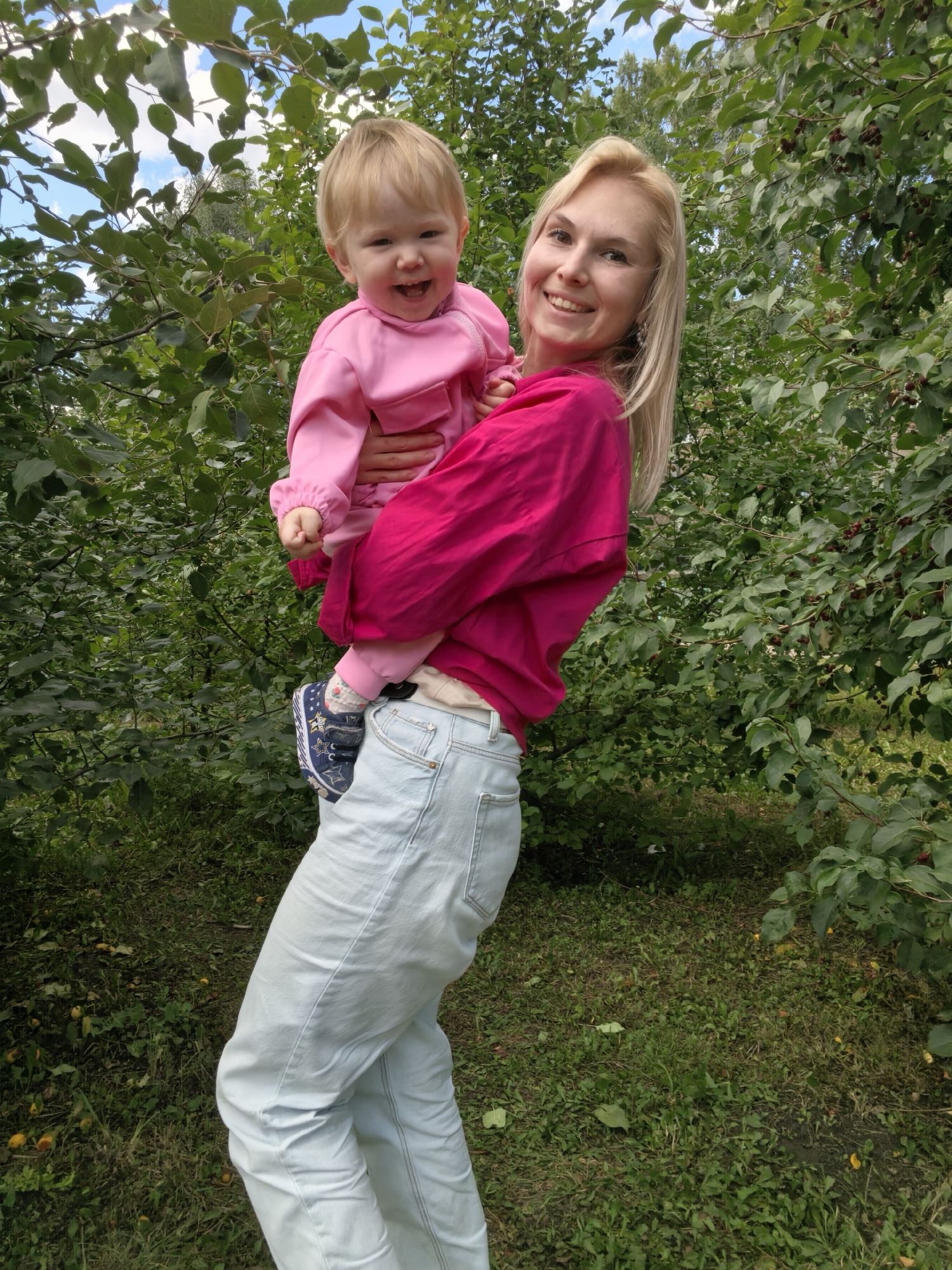 A blonde woman in a bright pink top and light blue jeans holds a young child, also in pink, in her arms outdoors. The child is smiling widely. They are standing in front of lush green foliage, with some dark berries visible on the branches. The ground beneath them is covered in grass and scattered yellow objects, possibly fallen fruit or leaves. The scene appears to be a park or garden in Sterlitamak, Russia, given the commonality of fruit-bearing trees and green spaces in the region. The sunlight suggests it is daytime.