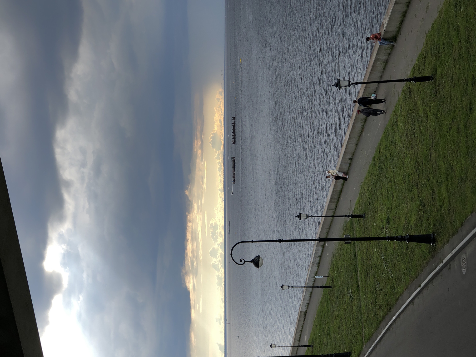 The image captures a view of a wide body of water under a dramatic, cloudy sky. In the foreground, a paved walkway runs alongside a grassy embankment. Several ornate black lampposts are spaced along the walkway. A concrete wall separates the walkway from the water.
Several individuals are visible on the walkway and near the water's edge. Two people are walking together on the walkway, and another person is sitting on the wall. Further along the walkway, two more people are standing. In the distance, on the water, are various boats, including what appear to be barges or small piers, and some smaller vessels. A faint line of distant land or structures is visible on the horizon. The sky is a mix of dark, heavy clouds and patches of bright, sunlit clouds, with rays of light breaking through in the distance. The water is choppy. The overall impression is of a public waterfront area, possibly a promenade or park, with a sense of impending or passing weather.
