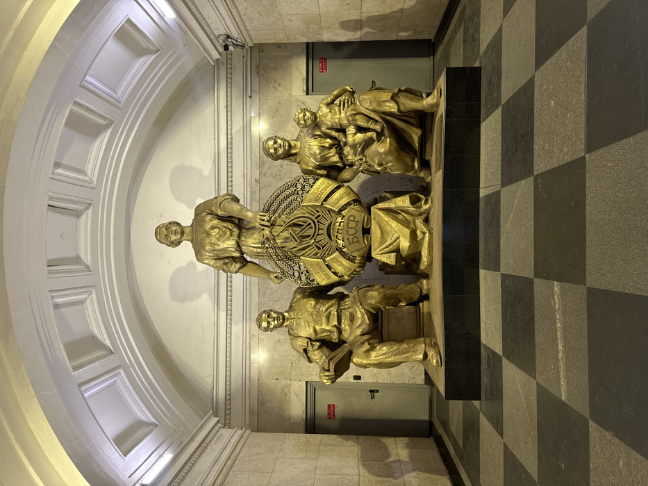 A wide-angle, eye-level photograph captures a monumental sculptural group located within Belorusskaya Station (Koltsevaya Line) of the Moscow Metro, Russia. The scene depicts a brightly lit, high-ceilinged underground hall.
The central subject is a large, golden-bronze, multi-figure sculptural composition positioned on a multi-tiered black plinth, facing directly towards the viewer. The group consists of four figures and a central emblem. From left to right: a seated male figure, rendered in golden-bronze, wearing work attire, holding an open book on his lap; a taller, standing female figure, also golden-bronze, positioned centrally and slightly elevated, holding a large circular emblem. This emblem is a golden depiction of the Coat of Arms of the Soviet Union, featuring the hammer and sickle, a globe, and wheat sheaves, with the prominent letters "СССР" at the bottom. The emblem rests on a draped golden pedestal. To the right, a seated female figure, golden-bronze, holds a child on her lap. All figures are cast in a uniform golden-bronze finish and look forward.
The background wall behind the sculptures is clad in polished light-colored stone, exhibiting horizontal decorative banding and subtle vertical elements. The arched ceiling above is painted white with recessed square panels and ornate molding. Two light-grey recessed doorways, each with a dark handle and a small red sign, are visible on either side of the sculptural group. The foreground floor is a checkerboard pattern composed of alternating dark grey and light grey polished square tiles. The scene is evenly illuminated, with light sources from above casting subtle shadows behind the statues on the background wall.