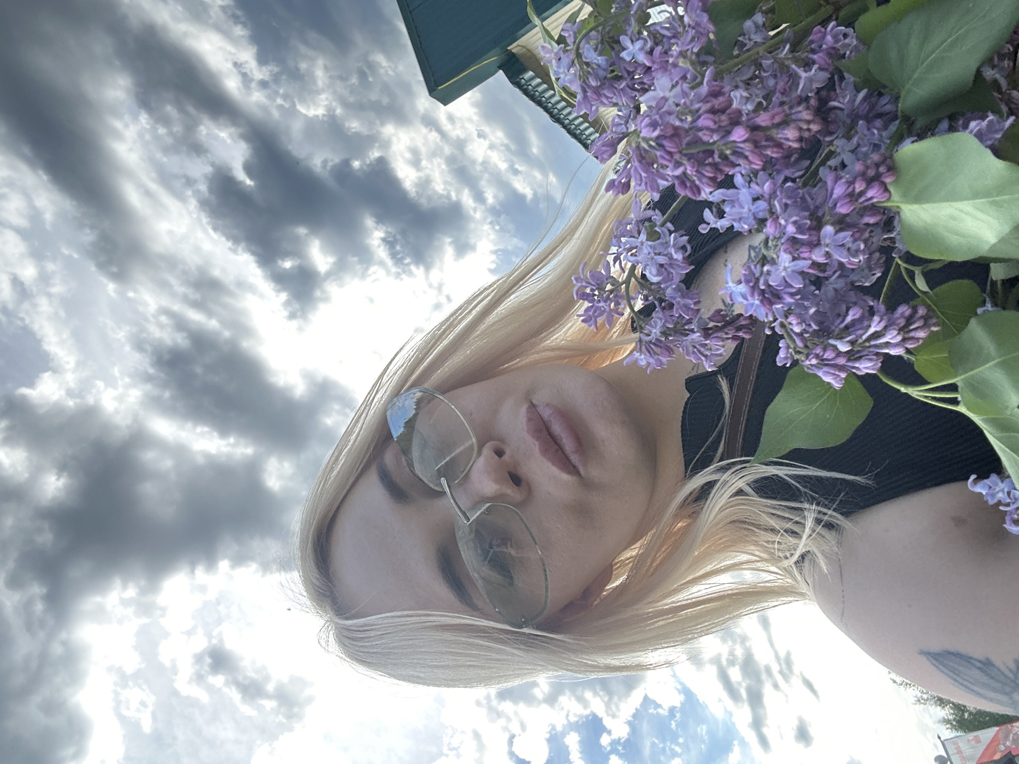 A low-angle selfie captures a blonde-haired woman holding a bouquet of purple lilacs outdoors. The woman, positioned centrally, wears sunglasses with light-tinted lenses that reflect parts of the sky and indistinct background elements. Her lips are slightly pursed, and a faint tattoo resembling a feather or leaf is visible on her upper left arm. She is wearing a dark, ribbed, sleeveless top. The bouquet of vibrant purple lilac flowers, with green leaves, is held in the lower right quadrant of the image. The background is dominated by a dramatic, partly cloudy sky, featuring large grey clouds interspersed with bright patches of sunlight. To the upper right, a dark green building with a visible roofline is partially visible against the sky. The lower left background shows very blurry, indistinct elements, possibly distant trees or structures. The scene is illuminated by strong natural light from above. The location is in the city of Unknown, Unknown.