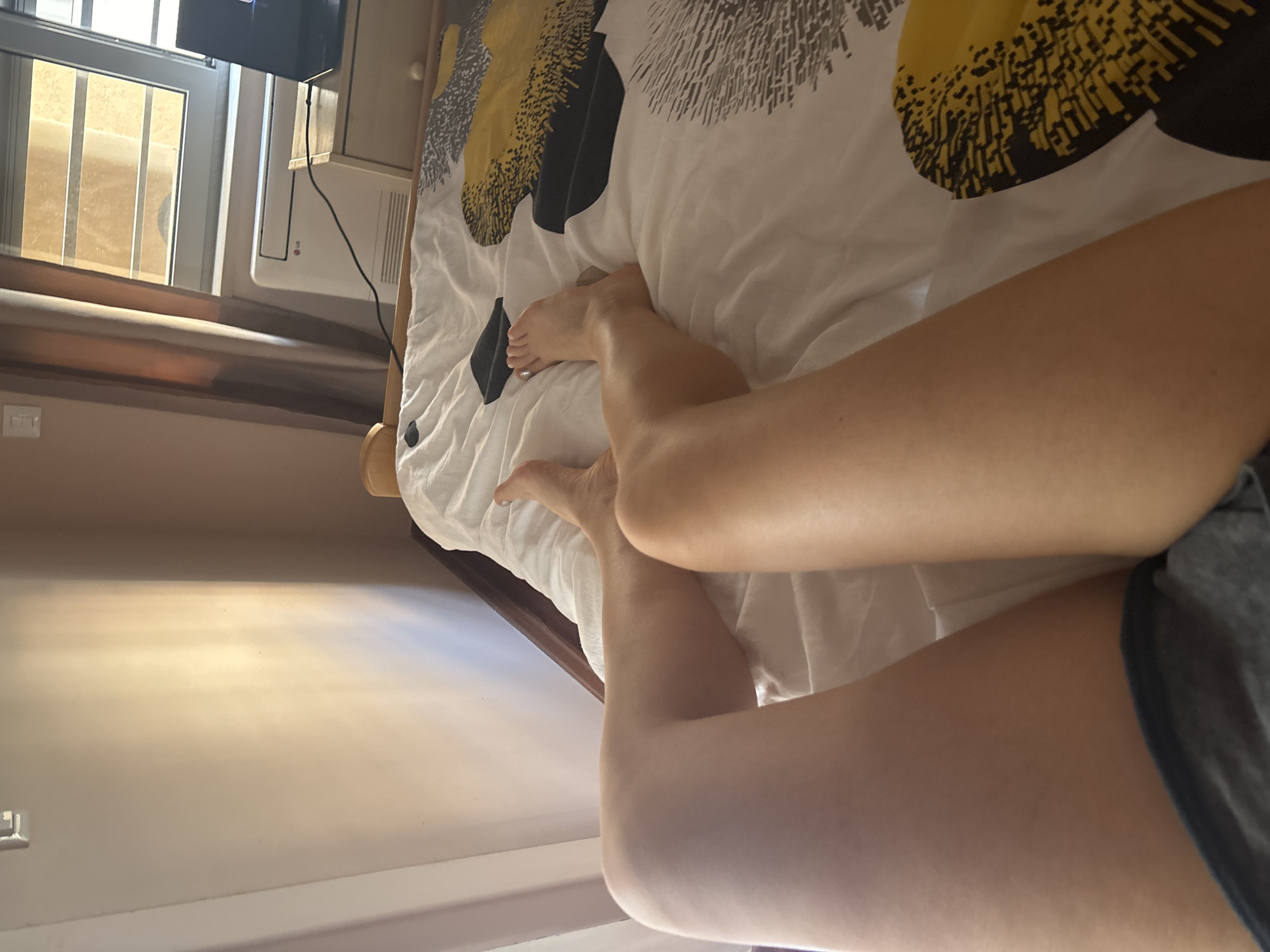 The image shows a person lying on a bed with a yellow and black patterned duvet. The person's legs are visible, with bare feet resting on the bed. The background includes a window with blinds, a wall-mounted air conditioning unit, and a beige wall. The location is likely an indoor space, possibly a hotel or apartment, given the presence of the air conditioning unit and the generic bedding. Budva, Montenegro, is not explicitly visible in the photograph.