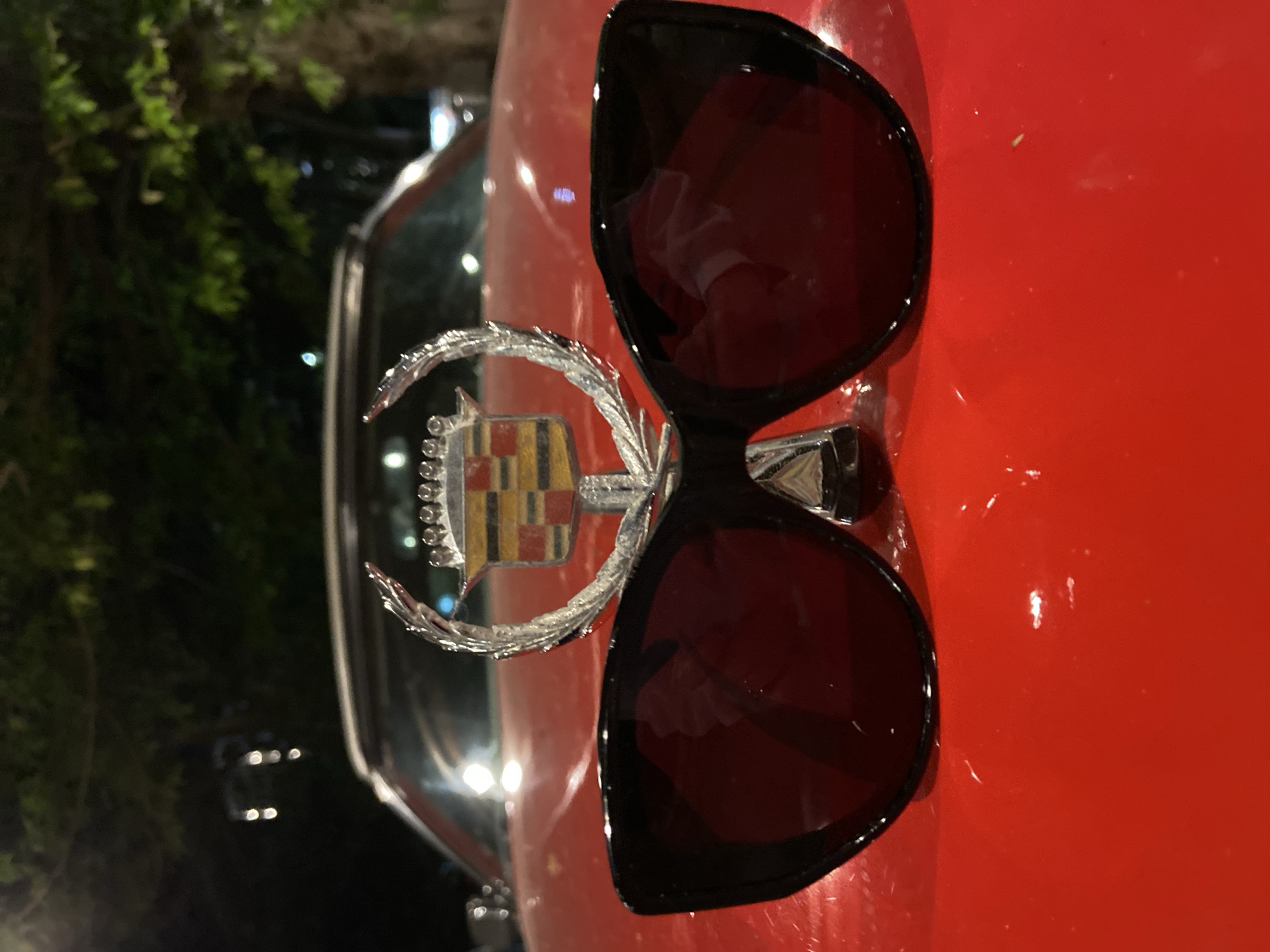The image features a close-up of the hood of a red car. Resting on the hood are a pair of black-framed sunglasses and a Cadillac hood ornament. The left side of the frame includes parts of the car's windshield and other dark elements, possibly foliage, suggesting an outdoor setting. The red paint of the car hood is smooth and reflects light. The hood ornament is chrome, depicting the Cadillac emblem with intricate details. The image does not depict Budva, Montenegro.