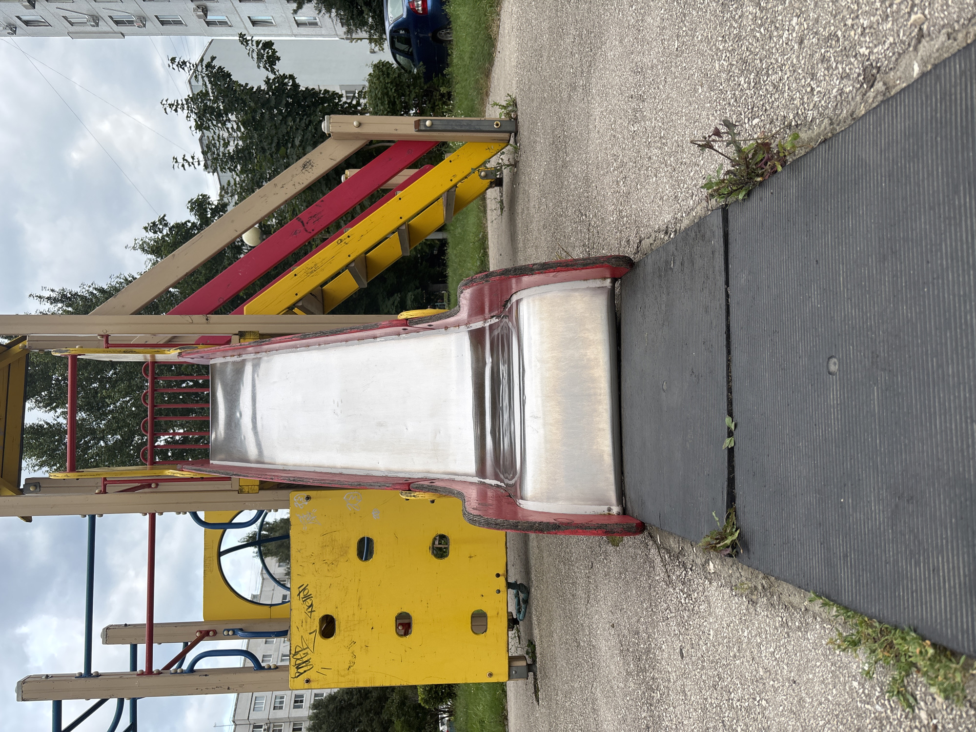 A low-angle, eye-level shot shows a section of a children's playground slide made of metal and red plastic trim, positioned on a black rubber mat. The slide extends upwards to a wooden structure with yellow and red-striped steps, and a yellow climbing wall with circular openings is visible to the left. Green grass and weeds grow around the edges of the playground. In the background, a blue car is parked near a tall, light-colored apartment building. The sky is overcast with clouds. The scene is located in Tolyatti, Russia.