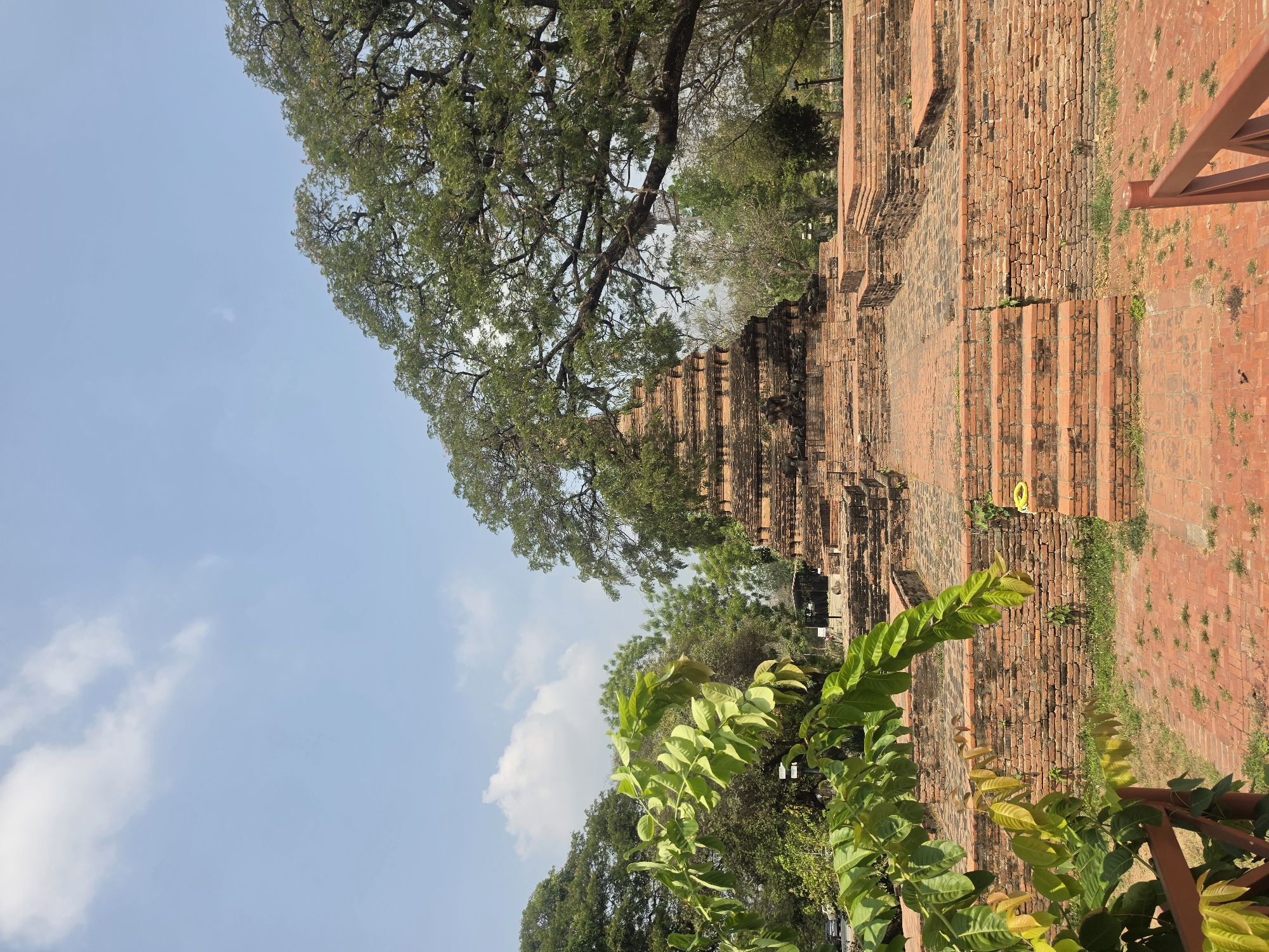 The image shows the ruins of Wat Yai Chaimongkol in the Historic City of Ayutthaya, Thailand. The main subject is a large brick stupa, partially surrounded by trees and greenery. In the foreground, lush green foliage is visible. The sky is light blue with scattered clouds. A portion of a red-orange metal structure is visible on the right side of the image. The image is taken from a low angle, looking up at the stupa.