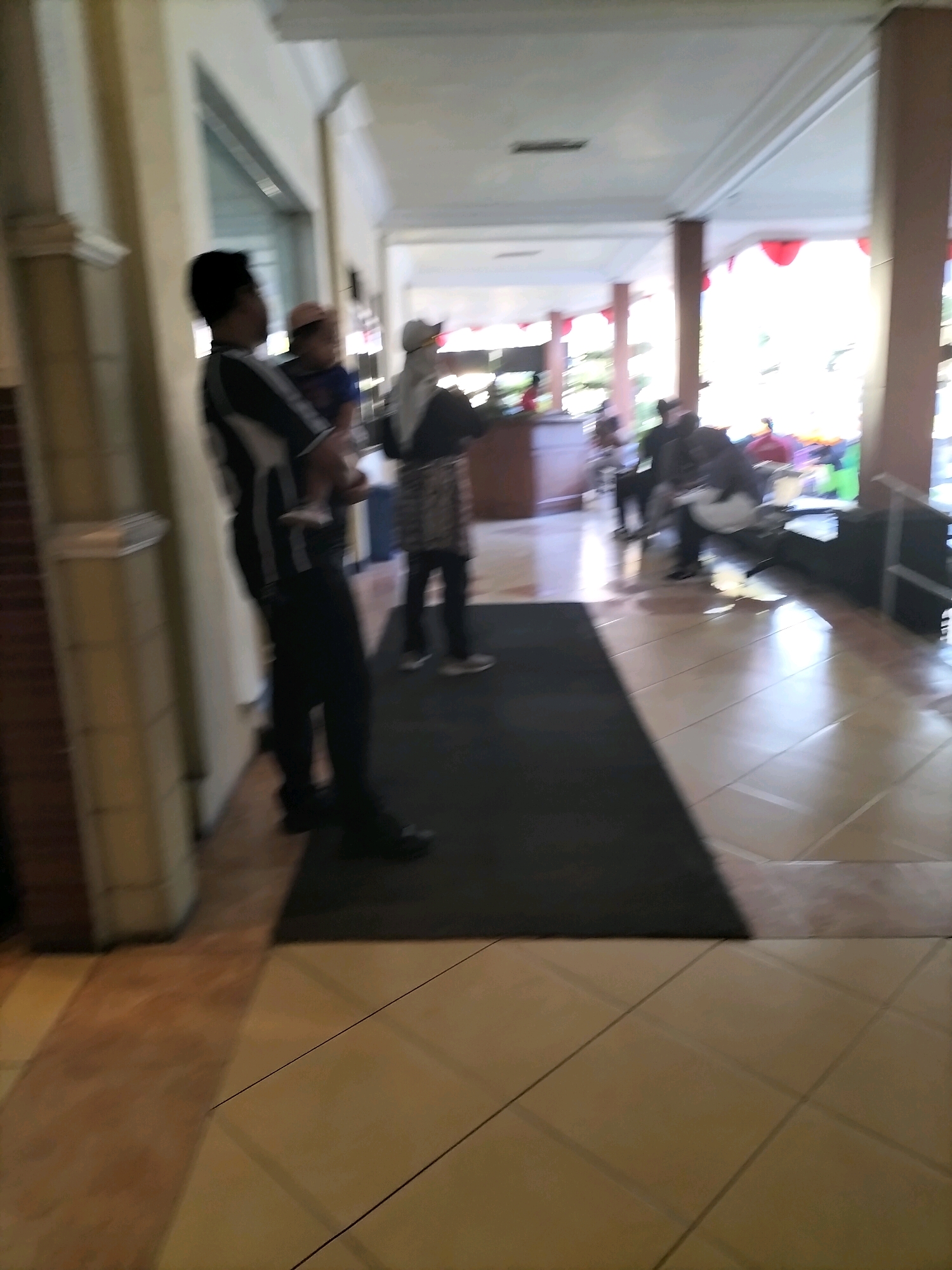 The image shows a group of people standing and walking in an indoor hallway. The hallway has tiled floors and a white ceiling with decorative molding. The walls appear to be a light color, possibly painted. In the foreground, a person in a black and white striped shirt and black pants is standing. To their left, another person is partially visible. Further down the hallway, two people are standing, one wearing a head covering. In the background, several other individuals are visible, some seated and others standing, suggesting a gathering or waiting area. Red decorative elements, possibly lanterns or fabric, are hanging from the ceiling in the mid-ground. The overall scene suggests a public indoor space, likely a hotel lobby, convention center, or a similar establishment in a warm climate given the apparent outdoor light entering the space. The location in Pare, Indonesia, is not explicitly indicated by any visual cues within the image itself.