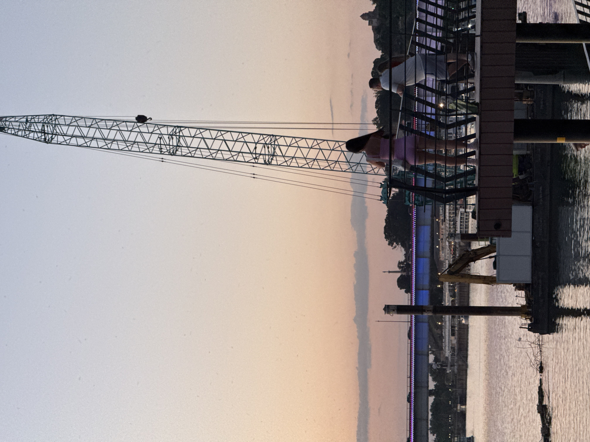 The image depicts a scene along the Sava River in Belgrade, Serbia, at dusk. In the foreground, a woman in a pink two-piece outfit stands on a boardwalk, facing away from the camera. Beside her, a man in a white polo shirt and grey shorts leans on the railing. Behind them, a large construction crane dominates the upper right portion of the frame, its metallic structure silhouetted against the twilight sky.
In the middle ground, the Sava River is visible, reflecting the soft colors of the sky and city lights. A pontoon with a small excavator and some debris is moored in the water near the foreground. Further across the river, a bridge illuminated with blue and purple lights spans the water, with cars visible on its surface. Beyond the bridge, the Belgrade Fortress (Kalemegdan) can be seen on the hill in the distance, its silhouette partially obscured by trees and buildings. The overall atmosphere is serene, capturing a moment of calm as day transitions into night by the river.