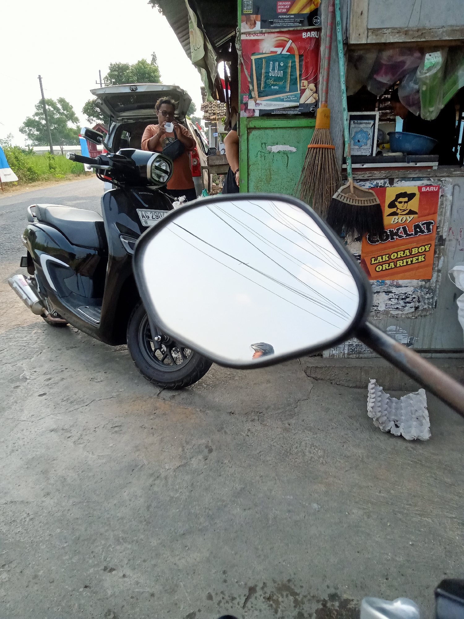 In the city of Bungur, Indonesia, a black Honda scooter is parked on a concrete surface near an open-air stall. A person, wearing an orange t-shirt and sunglasses, is standing by the open trunk of a car, holding a white cup and looking down at it. The stall displays various posters and items, including a broom and what appears to be cigarette advertisements. The scooter's side mirror reflects the sky and overhead wires, along with a glimpse of another person wearing a helmet. The environment appears to be an outdoor, possibly rural or semi-urban setting, with vegetation visible in the background.