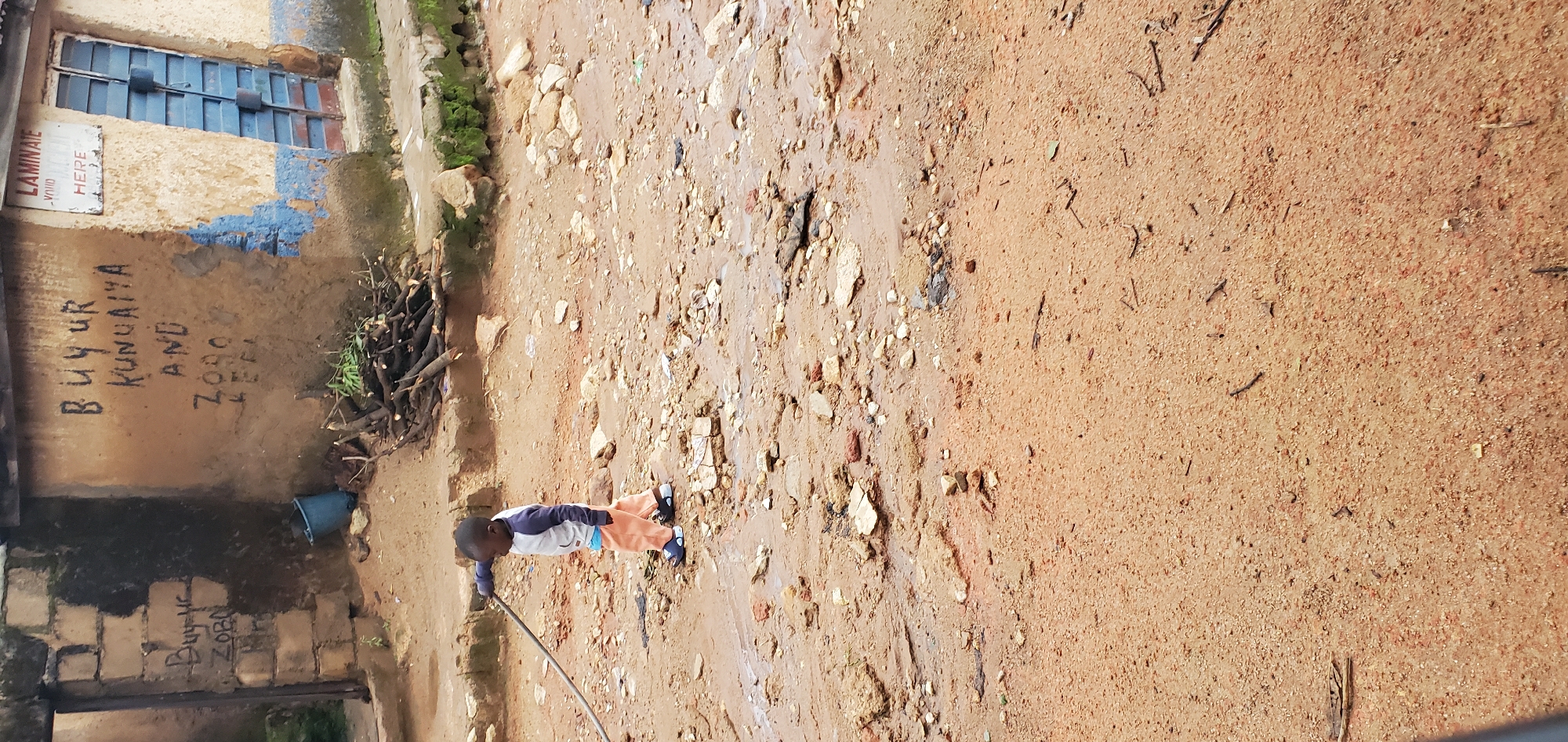 A young Black child, dressed in a gray and blue long-sleeved shirt and orange pants, walks on a dirt path towards the left of the frame. The child is holding a dark pole or stick. The ground is wet and muddy, with scattered rocks and debris. In the background, a weathered building stands with graffiti on its walls. One wall has black lettering that appears to read "BUY UR KUNUAIYA AND ZORO KEFI". To the right of this wall is a door with blue metal shutters. A sign above the door reads "LAMINATE YOUR HERE". A pile of cut branches is visible near the base of the building.