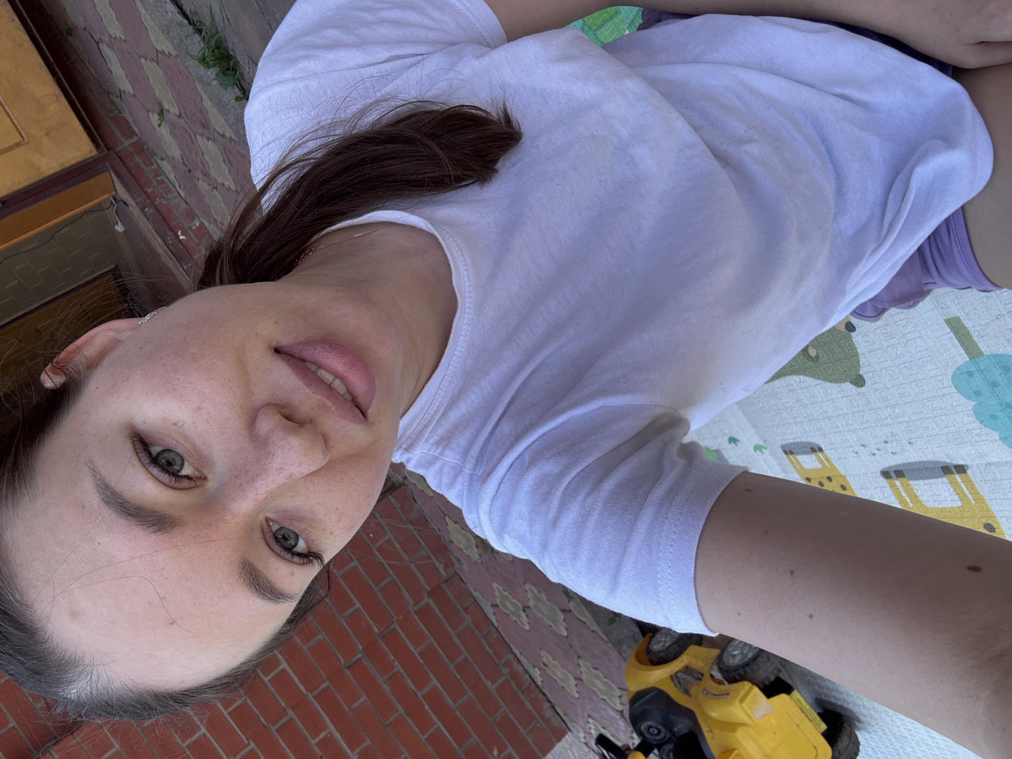 A woman is taking a selfie outdoors in Ферма, Russia. She is wearing a white t-shirt and purple shorts. Her hair is brown and pulled back. She is situated near a brick walkway and a building with red brick. Beside her is a play mat with illustrations of animals. A yellow and black toy vehicle is on the ground near the mat.