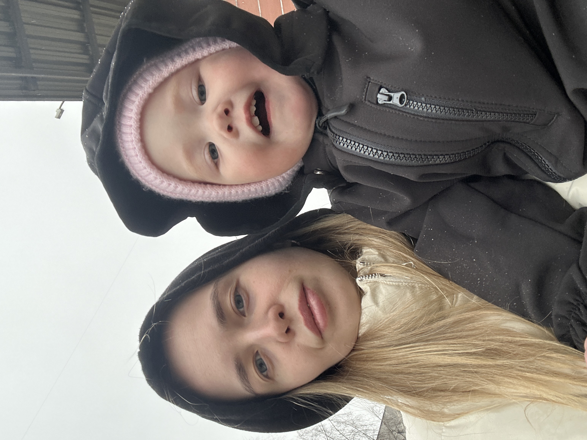 A woman and a toddler are posing for a selfie. The toddler is wearing a pink knit hat under a black hooded jacket, smiling with their mouth open showing some teeth. The woman is wearing a black hooded jacket and a cream-colored jacket underneath. She has light-colored hair. The background appears to be an overcast outdoor scene. Based on the context of the city of Chaikovsky, Russia, it may be a winter scene. Snow particles are visible on the clothing.