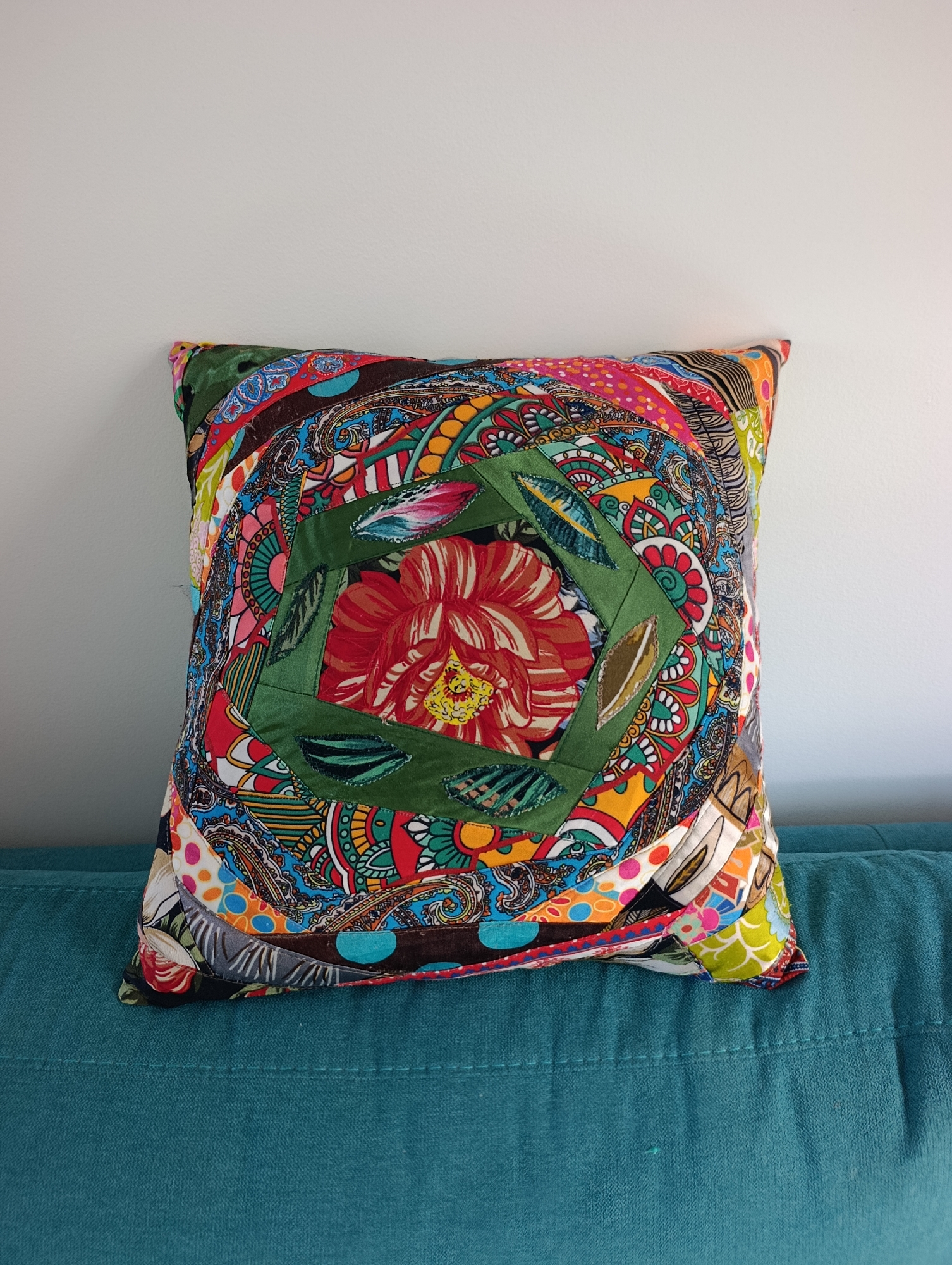 This is a close-up, eye-level, full shot of a brightly colored, square patchwork pillow with a swirling, paisley-like pattern. The pillow is placed on a teal-colored couch, and a white wall is visible in the background. The pillow's design features a central green square frame containing a red and yellow flower. The pillow's vibrant colors and intricate patterns are sharp and in focus, while the background is slightly blurred. The image does not contain any people or specific landmarks that would indicate its location in Setúbal, Portugal.