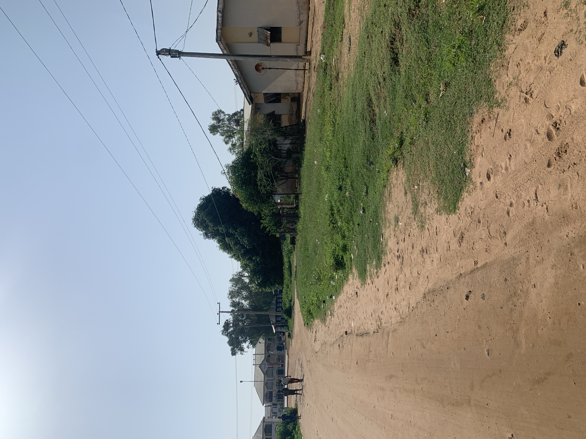 A vertical outdoor shot shows a dirt road with a grassy area to the left and a building on the far left. Several power lines stretch across the clear, bright sky. The road is light brown and has tire tracks and footprints. The grassy area is green and appears somewhat unkempt with some litter. A beige building with a metal roof and boarded-up windows is partially visible on the left. Further down the road, more buildings with pitched roofs can be seen, along with trees and utility poles. Two people are walking away from the camera on the road. The lighting suggests it is daytime, with a bright, clear sky. The overall scene appears to be a rural or semi-urban environment.