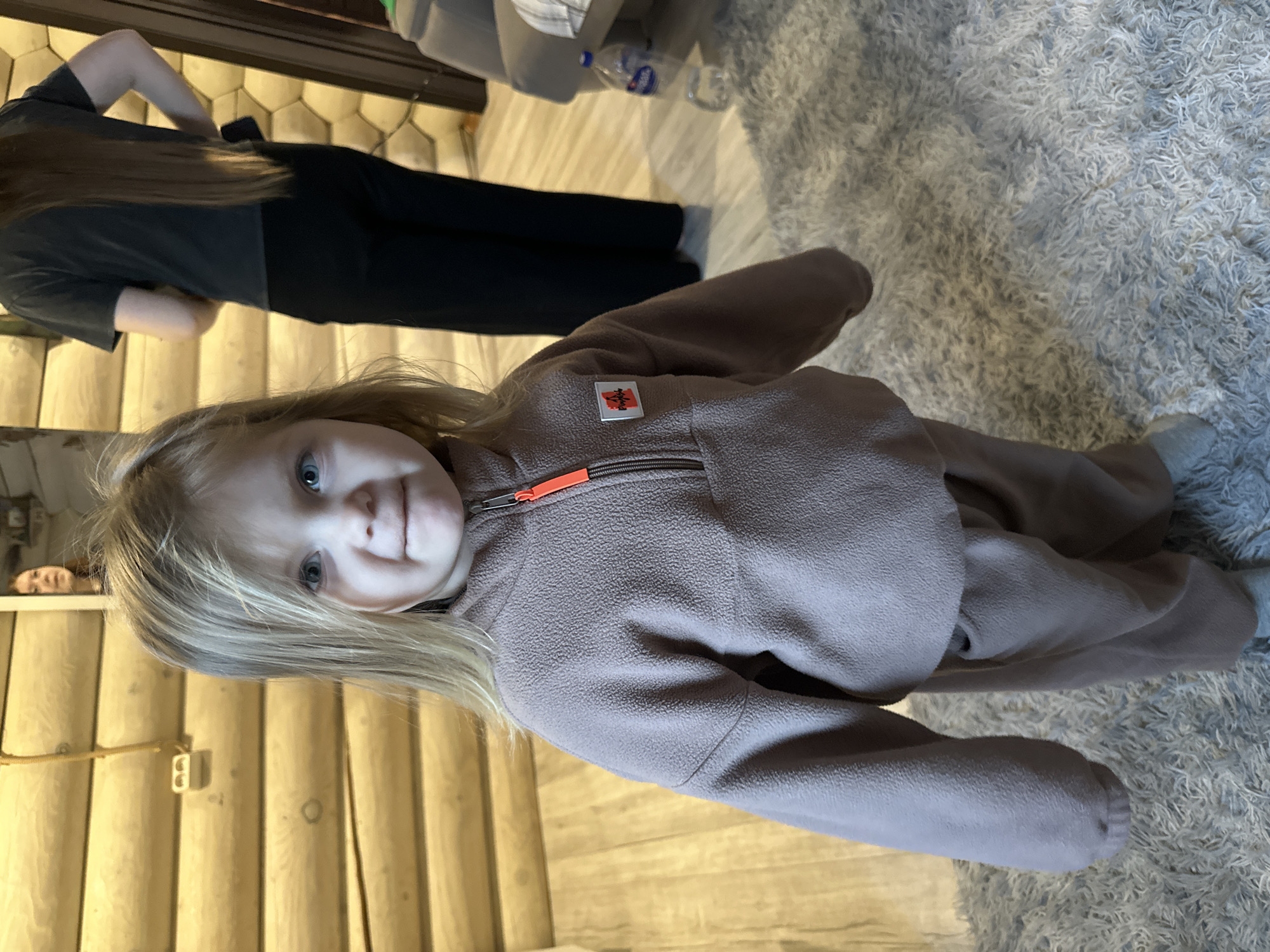 A young girl with blonde hair and blue eyes is standing indoors, wearing a mauve fleece jumpsuit with an orange zipper. In the background, an adult with long brown hair and black pants stands on wooden flooring with a light finish. The floor transitions to a gray shag carpet in the foreground. The interior has log walls. The location appears to be indoors, but information to verify the location in Ольховка, Russia, is insufficient in the provided picture.