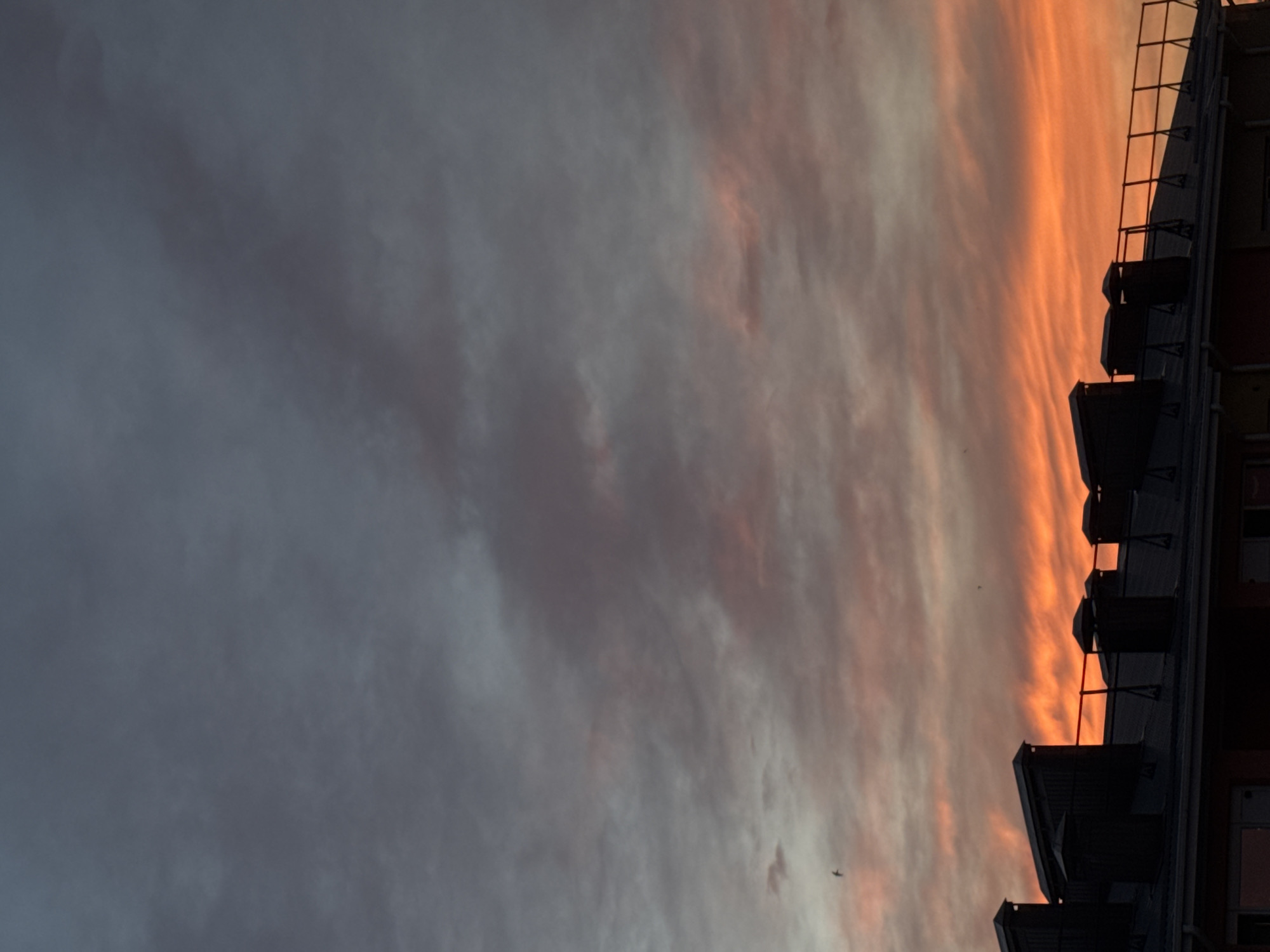 The image captures a rooftop scene at dusk in Mikhaylovsk, Russia. The sky is a dramatic gradient of dark grey at the top transitioning to vibrant orange and pink hues near the horizon, indicating the setting sun. Several chimneys, silhouetted against the colorful sky, stand prominently on the dark, angular rooftops of buildings. The rooftops themselves appear to be constructed of dark material, likely asphalt shingles or metal, and are lined with what seem to be ventilation or chimney structures. The overall impression is one of a calm, urban evening, with the natural beauty of the sunset contrasting with the man-made structures of the city.