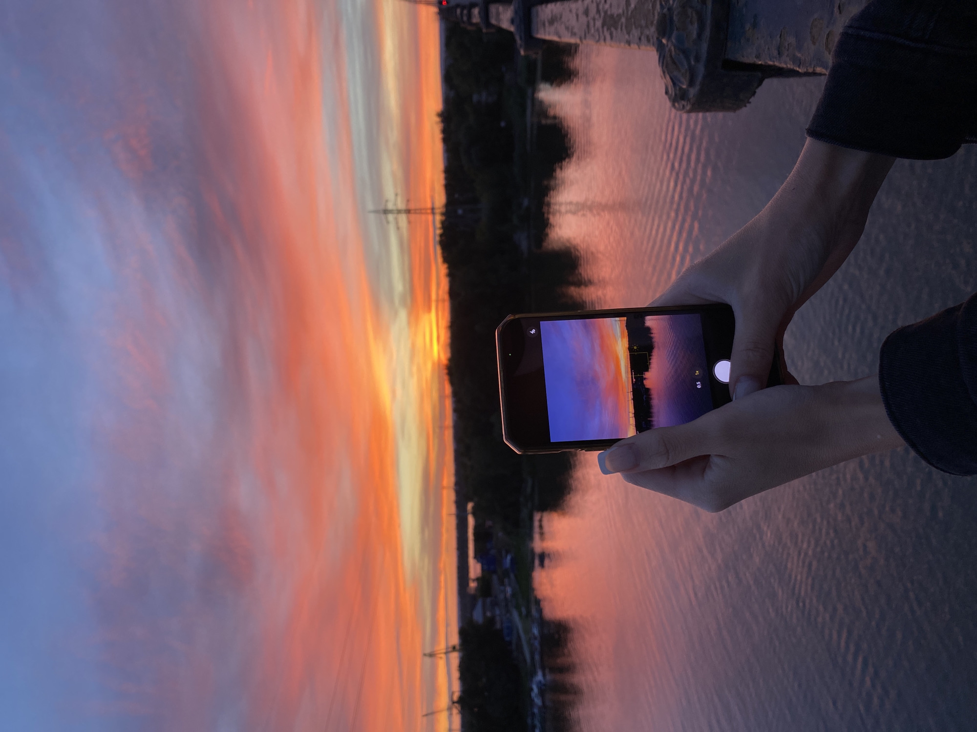 In an outdoor scene at sunset, a person's hands hold a smartphone, capturing a photograph of the vibrant sky and its reflection on a body of water. The sky displays a gradient of colors, transitioning from a soft blue at the top to shades of pink, orange, and yellow towards the horizon. Wispy clouds are scattered across the sky, enhancing the sunset's dramatic effect. The water below mirrors these colors, creating a picturesque scene. In the background, a treeline separates the water from a distant urban landscape featuring industrial cranes and utility poles. The foreground includes a bridge railing, suggesting the vantage point from which the photograph is taken. The smartphone screen shows a captured image of the sunset, with UI elements like zoom levels (0.5x, 1x) visible.