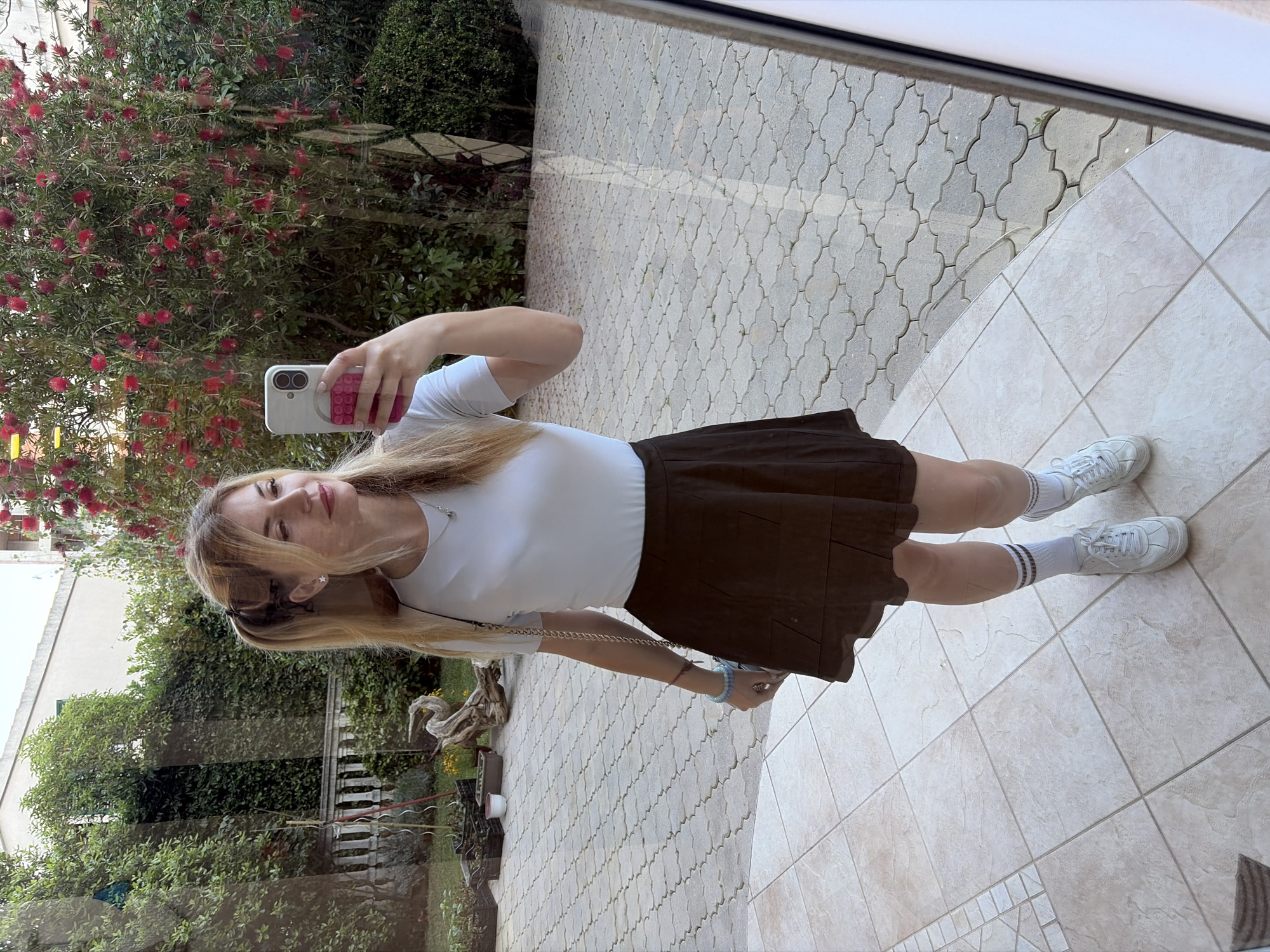 A young woman with long blonde hair is taking a selfie in a courtyard, reflected in a window. She is wearing a white short-sleeved shirt, a short brown skirt, white socks with black stripes, and white sneakers. Her phone has a white case with a pink design.
The courtyard is paved with patterned gray stones. In the reflection of the window, there is a garden with a variety of plants, including a large bush with red flowers. The setting suggests an outdoor area in a residential or commercial district in Budva, Montenegro.