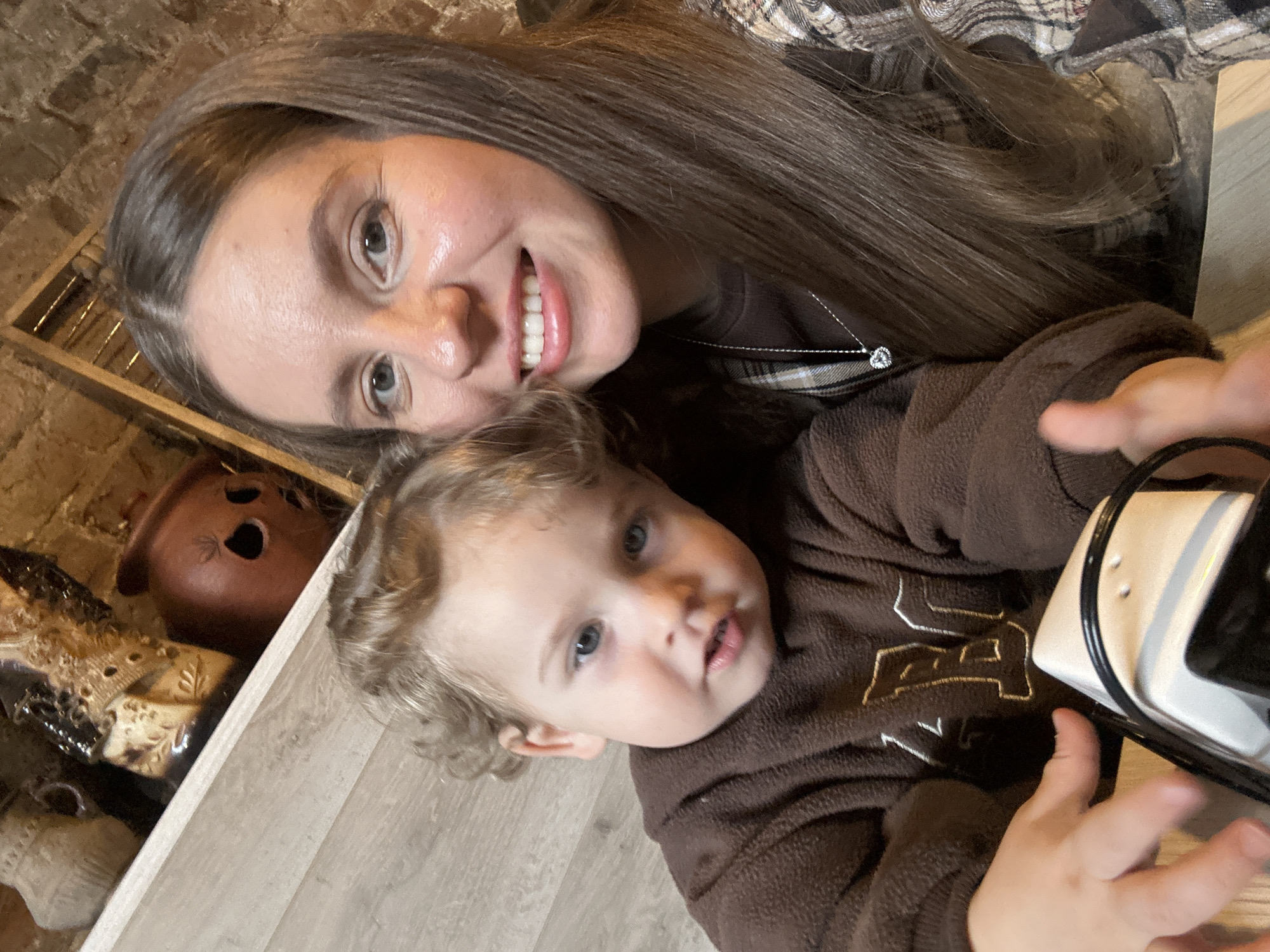 This image, captured indoors in Klin, Russia, features an adult female and a young child in a close-up, selfie-style portrait. The adult female, positioned on the right, has long brown hair parted in the middle and is smiling directly at the camera. She wears a dark brown long-sleeved top under a plaid shirt with brown, beige, and white checks, and a silver chain with a heart-shaped pendant is visible around her neck. The young child, situated on the left, appears to be between one and three years old, possessing curly light brown hair and blue eyes, looking towards the camera with a slightly open mouth. The child is wearing a brown fleece sweatshirt with a light-colored embroidered letter, possibly 'M' or 'N', on the chest. The child's right hand is visible in the lower left, positioned near a white and black object with metallic trim, possibly a toy or small electronic device. In the background, a light-colored wooden shelf displays several decorative items: a textured beige vase, an ornate dark-colored sculpture, and a reddish-brown, mask-like ceramic object with hollow eye sockets. Behind the shelf, an exposed brick wall is visible. The surface in the lower foreground is light wood-grain. The scene is illuminated by warm, likely artificial, lighting.
