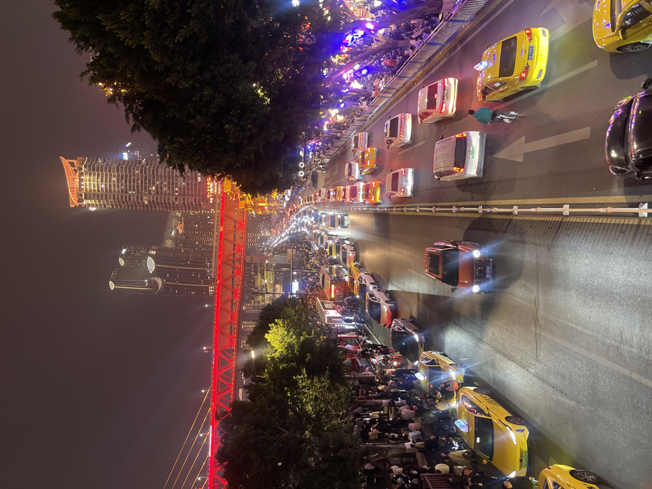 This nighttime image depicts a bustling urban street scene in Yuzhong District, Chongqing, China. The perspective is elevated, looking down upon a multi-lane roadway filled with a high volume of vehicles and flanked by dense crowds of pedestrians.
The road, illuminated by streetlights and vehicle headlights/taillights, shows heavy traffic, primarily consisting of cars, many of which are yellow taxis, suggesting congestion or slow movement. An orange SUV is visible in the foreground, with numerous other vehicles, including a silver sedan, white sedans, and more yellow taxis, extending into the distance. A lone individual on a bicycle, wearing a teal-colored jacket, is observed in the right-center lane, cycling away from the viewer. Lane markings, including double yellow lines and white dashed lines, are visible, along with a prominent white arrow painted on the pavement indicating the direction of travel.
On both sides of the street, large groups of pedestrians are present. On the left, a dense crowd occupies a sidewalk adjacent to commercial establishments and a bus stop. On the right, pedestrians are gathered behind a metal barrier, beneath trees adorned with purple string lights, suggesting a festive or commercial atmosphere.
In the mid-ground, lush green trees line both sides of the thoroughfare. In the background, the prominent Dongshuimen Bridge, illuminated in a vivid red, spans across the scene. Beyond the bridge and to its right, a cluster of modern high-rise buildings and skyscrapers dominates the skyline, brightly lit against the dark night sky. One skyscraper on the right features a distinctive illuminated crown. The overall environment is one of an active, densely populated urban area, brightly lit at night.