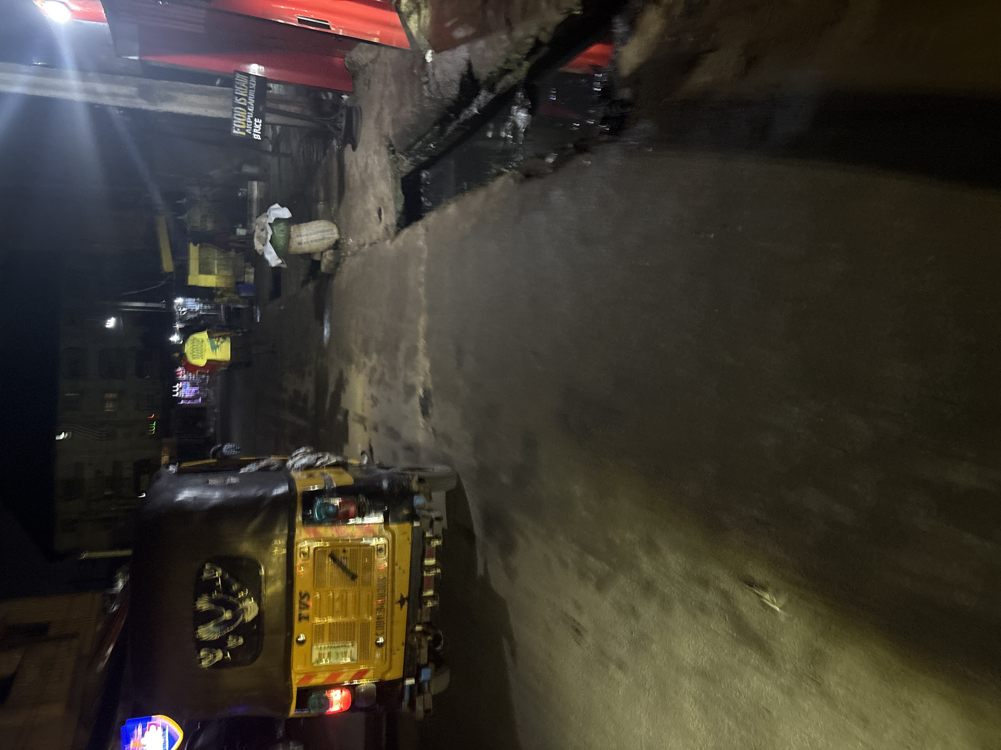 A yellow auto-rickshaw with an illuminated sign reading "TVS" is parked on a street in Onitsha, Nigeria. The auto-rickshaw has decorative designs of birds on its rear window. The street is wet, suggesting recent rain, and a drainage channel runs alongside the road. A sign on a pole reads "FOOD IS READY AKPU GARRISER PRICE". Several people are visible in the background, including one wearing a yellow t-shirt. The scene is illuminated by streetlights and neon signs, indicating it is nighttime.