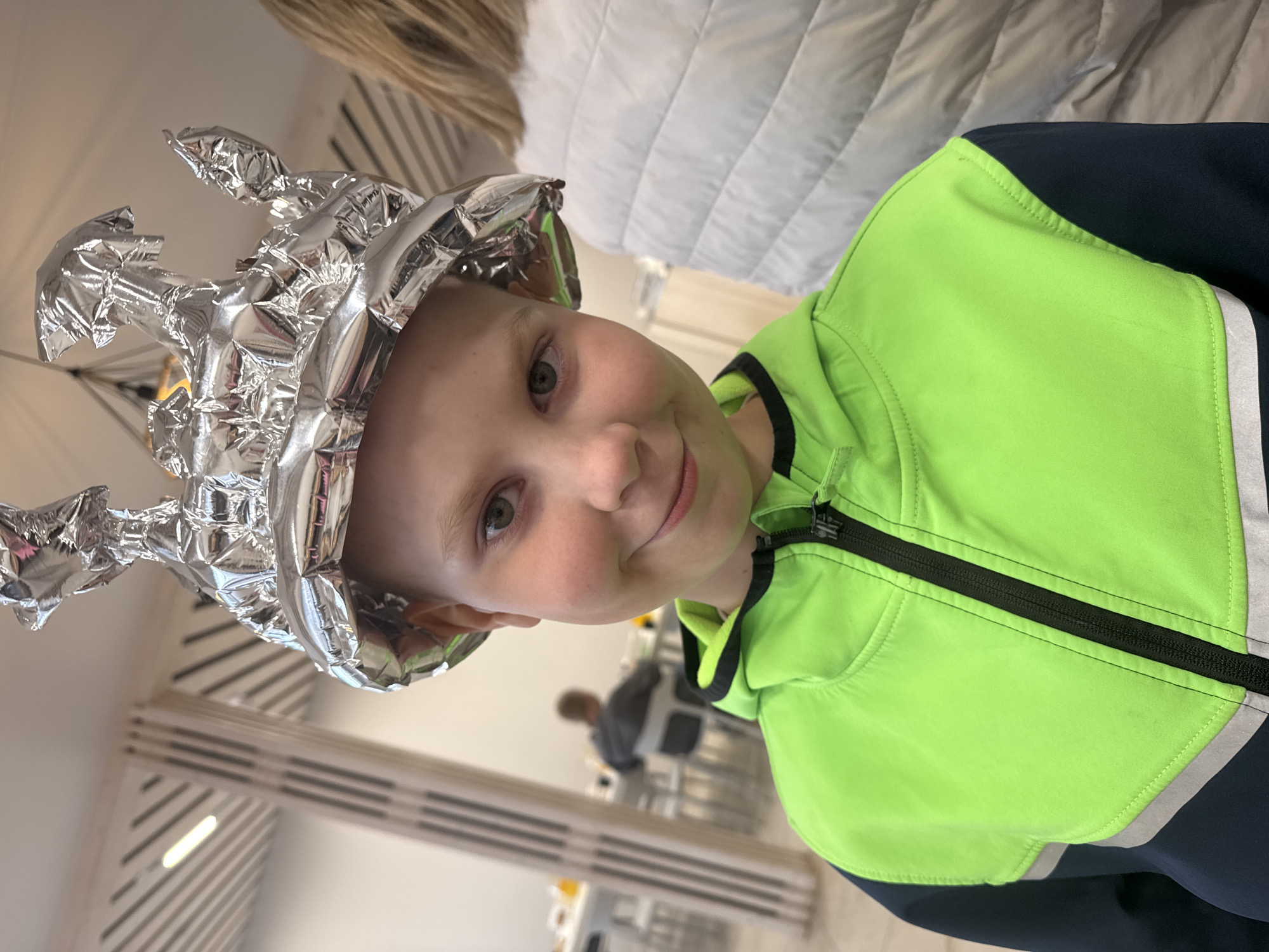 A child is indoors wearing a makeshift hat constructed from aluminum foil. The hat is irregularly shaped with pointed extensions. The child is wearing a bright green and dark blue jacket with a black zipper. The background is blurred, but suggests a public space with tables and chairs. The location is in the city of Chaikovsky, Russia. Other individuals are visible in the background.