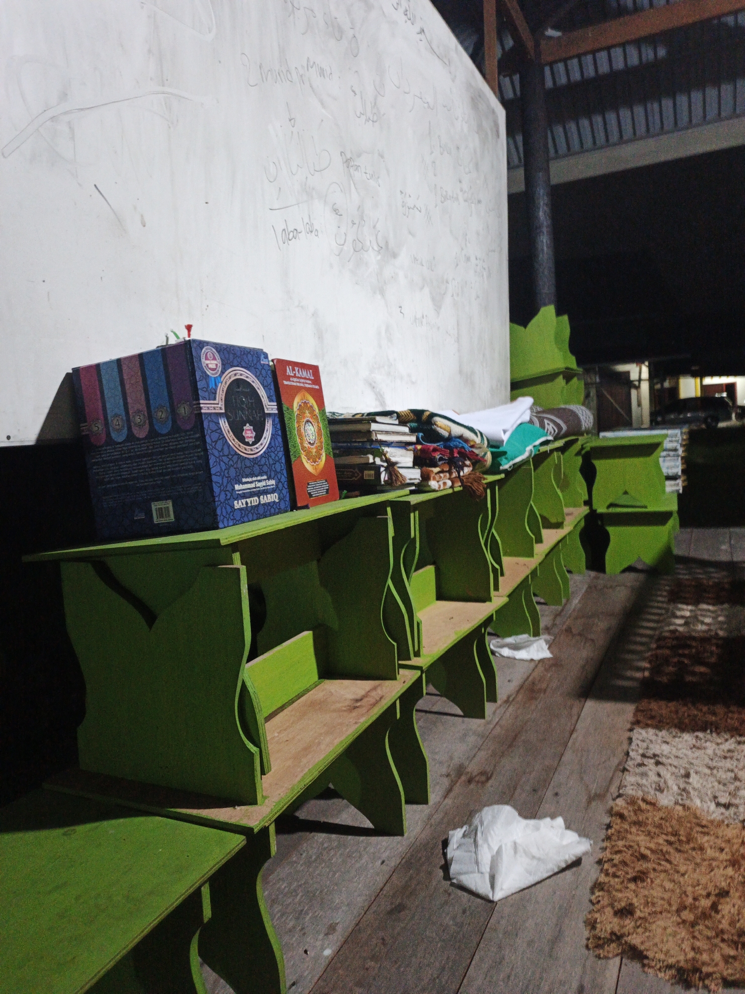 The image shows an indoor scene with a white wall on the left, covered in handwritten Arabic script. In front of the wall, there are several green wooden shelves, some of which are empty and others contain books and other items. On the top shelf, two prominent books are visible: a large blue and black book with gold lettering that reads "FIQH SUNNAH" and a smaller orange and yellow book with Arabic text. Stacked behind these are more books and some folded fabric, possibly prayer rugs. The shelves are arranged in a line, receding into the background. The floor is made of wooden planks, and a textured rug is visible on the right side of the frame. The overall lighting suggests an indoor environment, possibly a place of worship or study. Based on the provided context, the location is identified as Kelapapati, Indonesia.