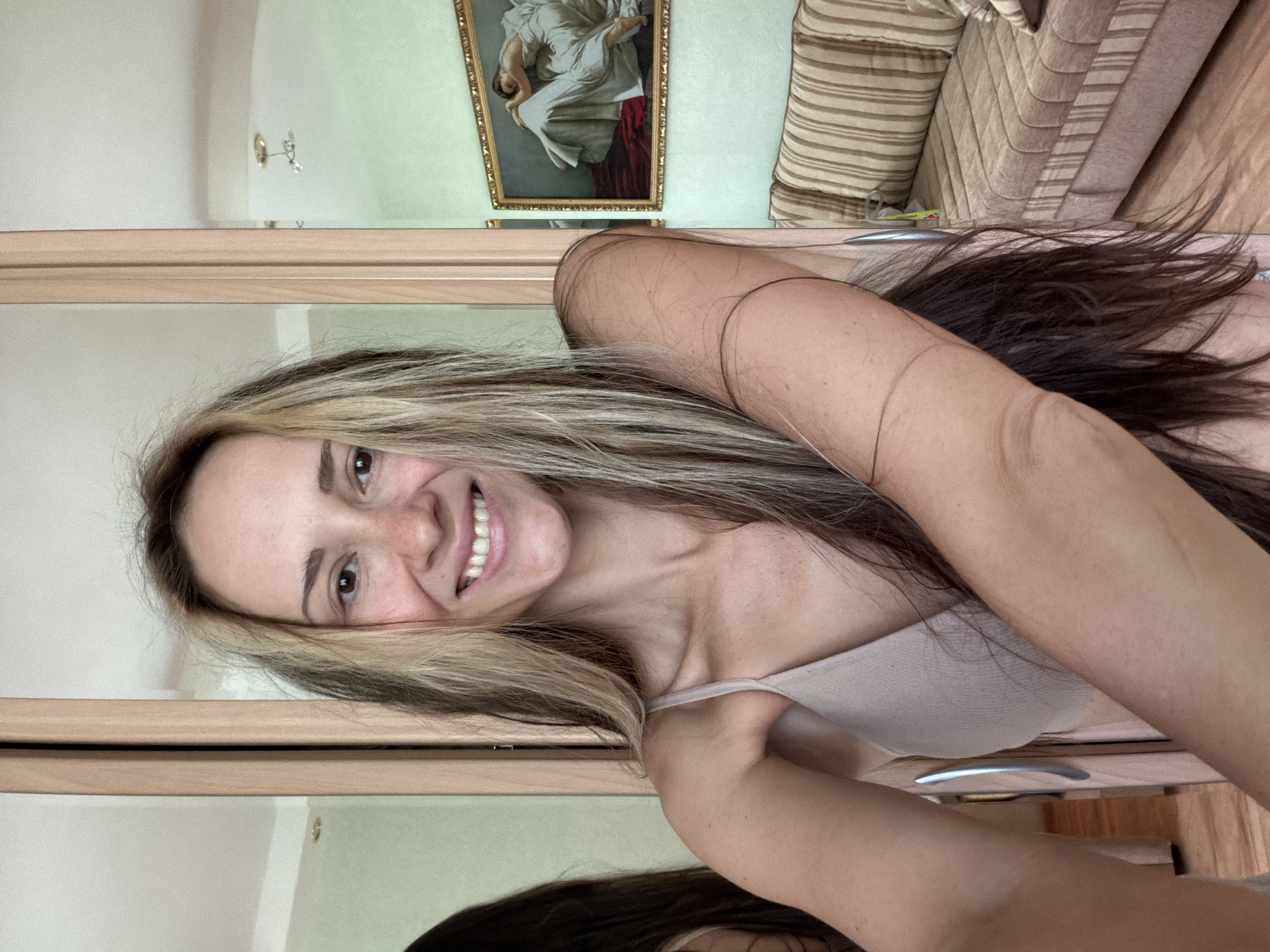 A woman with long, highlighted brown hair is smiling at the camera. She is wearing a light-colored tank top. A mirror reflects her image and the background, which includes a painting in a gold frame, a striped couch, and a light-colored wall. The location is Kamyshin, Russia.