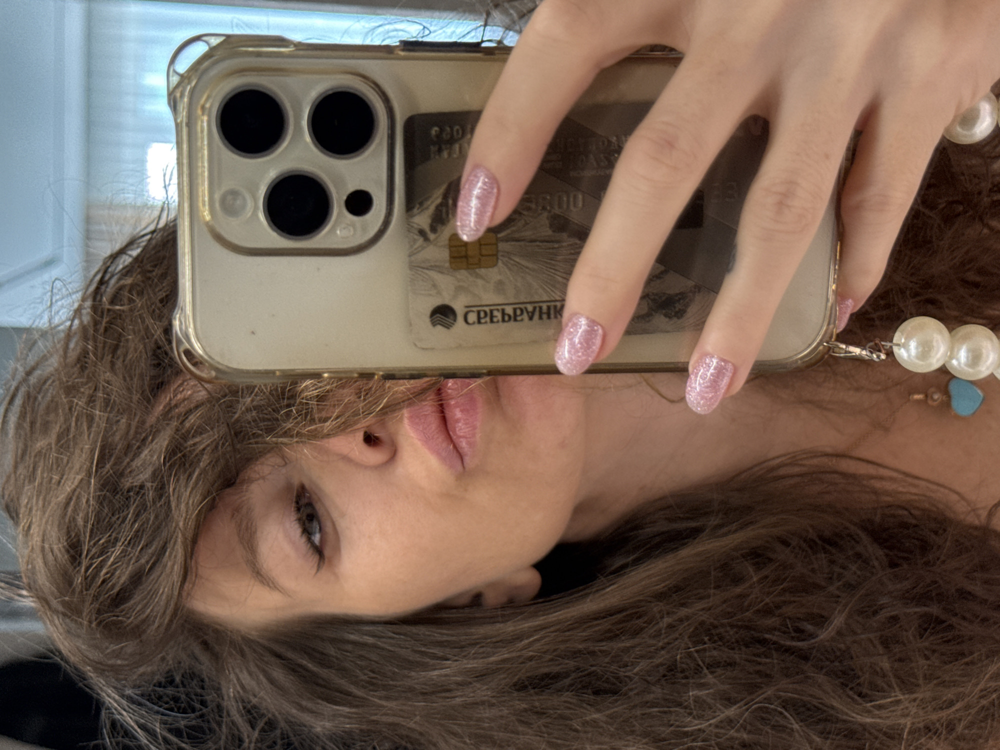 This inverted close-up selfie captures a person, likely female, in an indoor setting in Khimki, Russia. The primary subject's head is tilted sideways, with her dark brown, wavy hair framing her face. Her left eye, enhanced with eyeliner, and her subtly made-up, light pink lips forming a gentle pout, are visible.
A hand, adorned with short nails painted in sparkling pink glitter polish, holds a clear, slightly yellowed smartphone case. The smartphone features a prominent triple-lens camera system on its rear. Tucked into the phone case is a light-colored bank card, partially visible. On the card, the metallic chip is prominent, along with Cyrillic text that reads "СПЕРБАНК" (SBERBANK) and "МИР" (MIR), a Russian payment system. Other blurred numbers, including what appears to be "3800," are also visible on the card.
On the right side of the frame, a segment of a large, white pearl necklace is visible, featuring a small turquoise heart-shaped bead accent. The background is lightly blurred, suggesting an indoor environment with a bright area, possibly a window, on the left side of the frame. The overall impression is a casual, personal photograph.