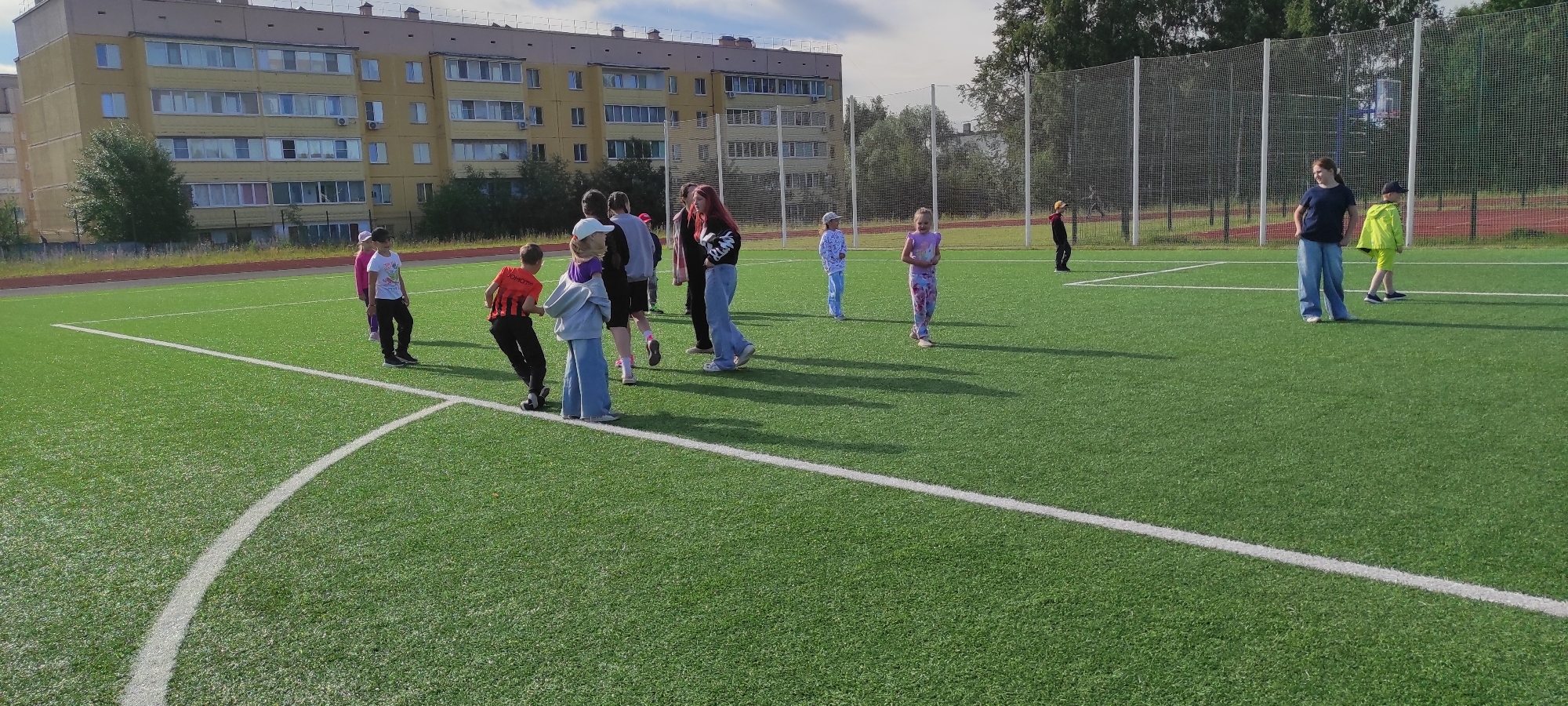 The image depicts an outdoor artificial turf field, likely a sports complex, in Noviy, Russia. A multi-story residential building with a light yellow facade, featuring numerous windows and balconies, is visible in the background to the left. Several children and young adults are scattered across the field, engaged in various activities. Some appear to be observing or interacting with each other, while others are standing or walking. A chain-link fence with white poles encloses the field in the background, and trees are visible beyond the fence. The field is marked with white lines, indicating boundaries or segments of a sports area. The sky is partly cloudy.
