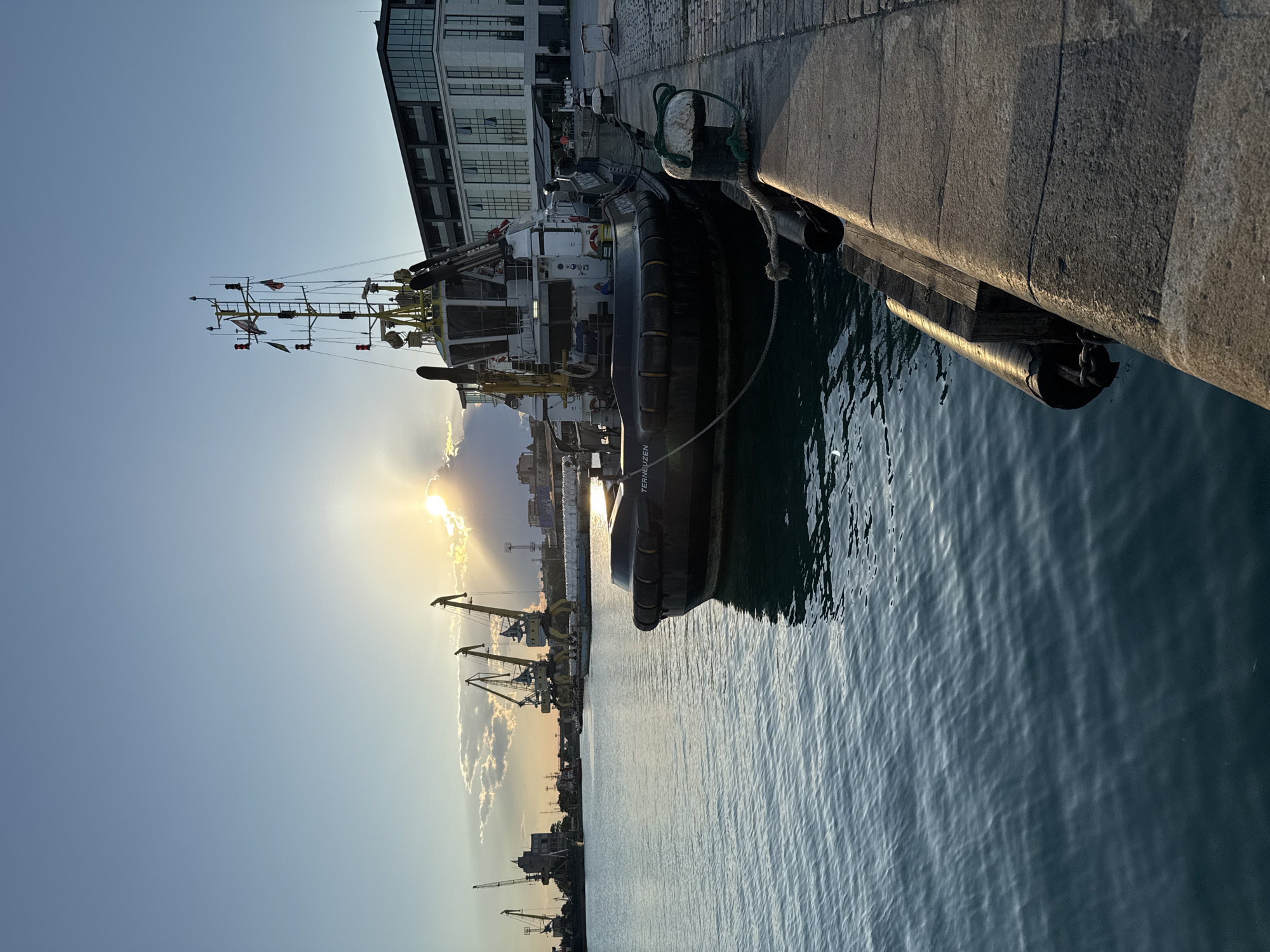 The image depicts a harbor scene in Burgas, Bulgaria, during sunset. A tugboat is moored to a stone quay in the foreground, with its name "TERNEUZEN" visible. The water is calm, reflecting the sky and surrounding structures. In the background, several large cranes stand silhouetted against the setting sun, indicating industrial activity. A modern building with large glass windows is visible to the right of the tugboat. The sky is a blend of blue and orange hues, with clouds catching the sunlight. The overall atmosphere is serene and industrial.