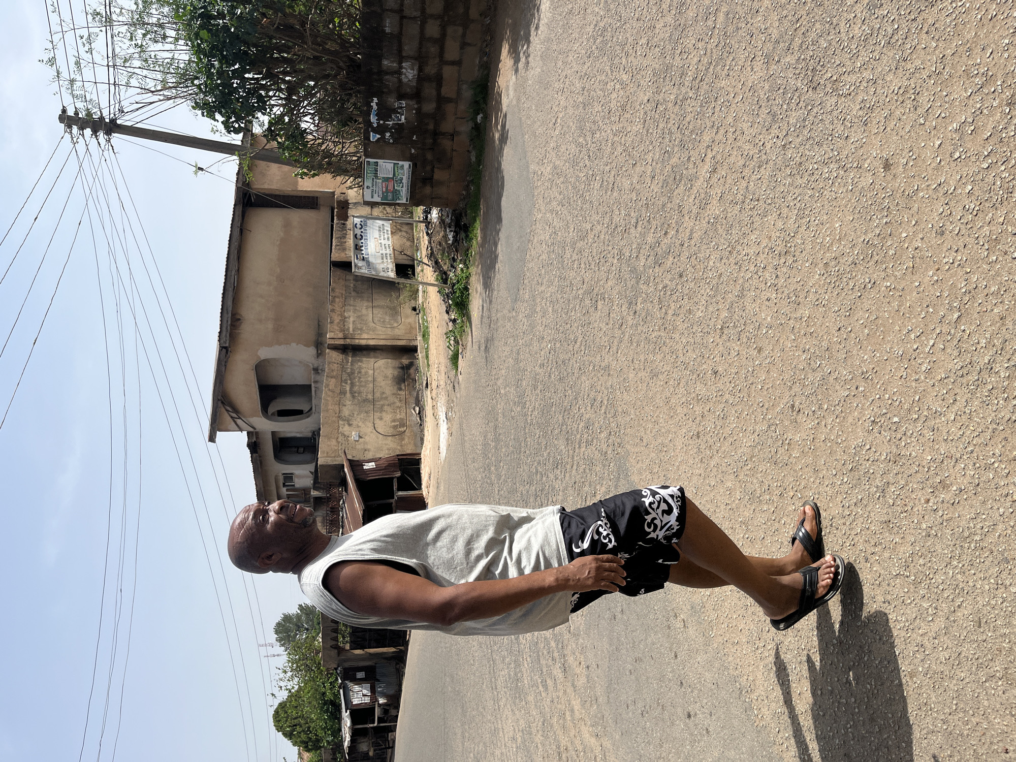 A medium shot, taken at eye level, portrays a dark-skinned man walking on an asphalt road in Jos, Nigeria. The man is bald and is wearing a light gray sleeveless shirt and black shorts with white designs. His footwear consists of black sandals. He is looking slightly upward and to the left, possibly observing something of interest.
The road surface is rough, with a mix of fine and coarse aggregates. To the left of the man are buildings constructed of concrete and brick, with some sections showing signs of weathering and disrepair. Power lines stretch across the sky, supported by a utility pole.
Vegetation, including trees and bushes, is visible along the edge of the road and behind the buildings, indicating the urban landscape.