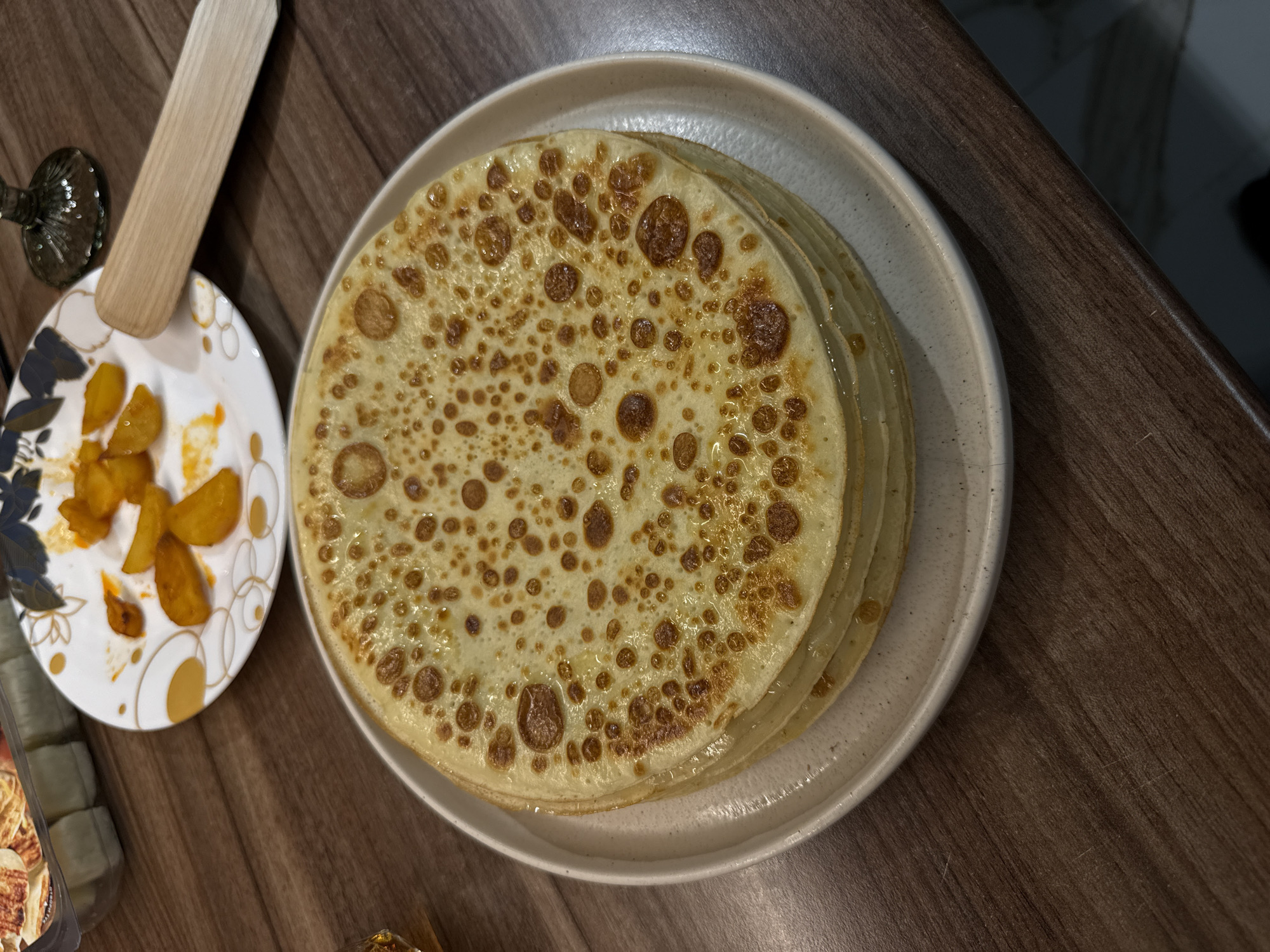 The image depicts a stack of thin pancakes, likely crepes, on a round, light-colored plate. A smaller plate to the left contains what appears to be pieces of cooked potatoes or a similar food item, and a wooden spatula is resting on this plate. The scene is set on a dark brown wooden surface, likely a tabletop. The lighting is soft and warm. There is insufficient information to identify a specific location in Sofia, Bulgaria.