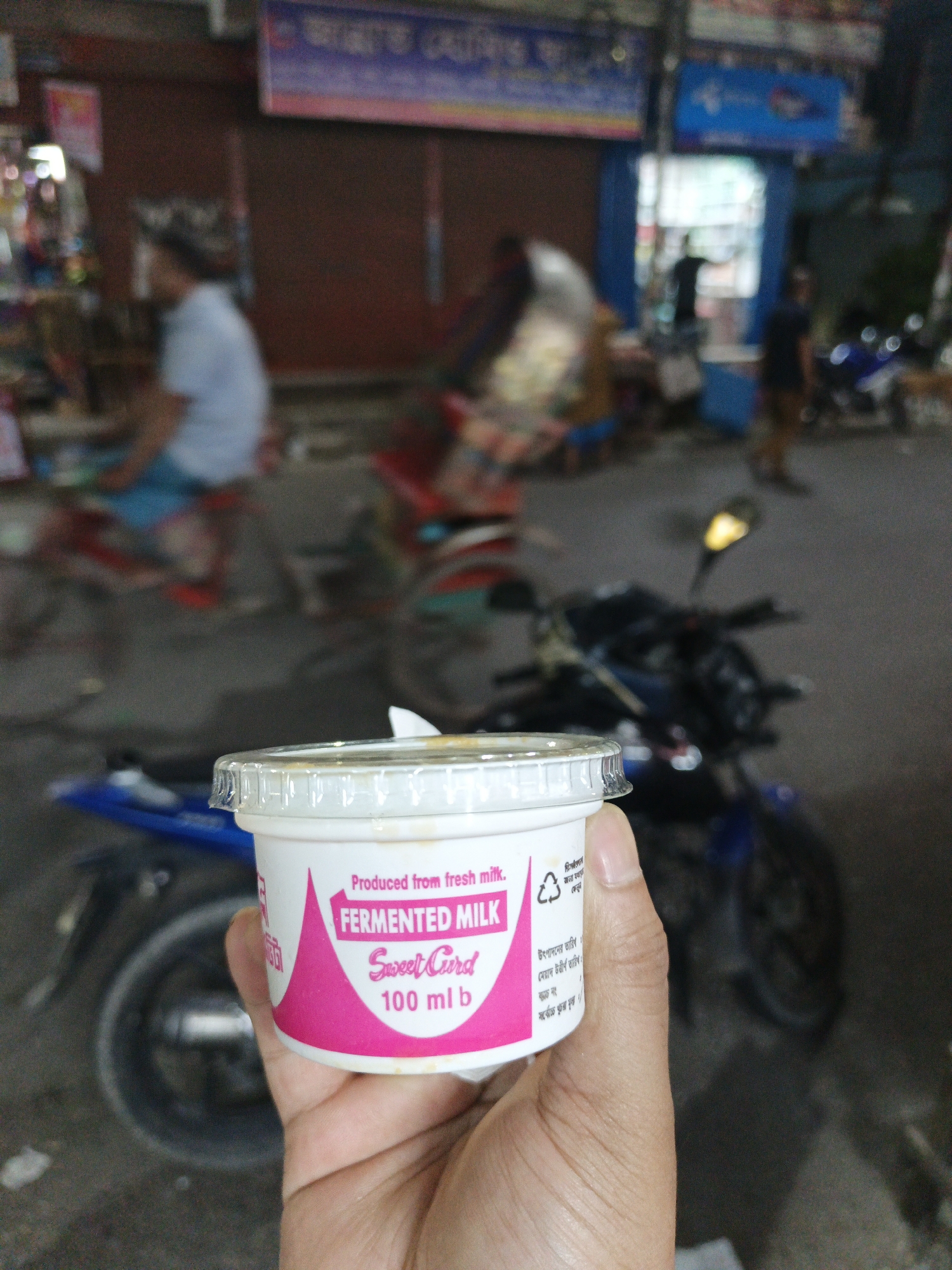 A person's hand holds a container of "Sweet Curd" fermented milk. The container is white with pink labeling, indicating it's produced from fresh milk and contains 100 ml. In the blurry background, there's a street scene with a motorcycle, people, and buildings, suggesting an urban setting.