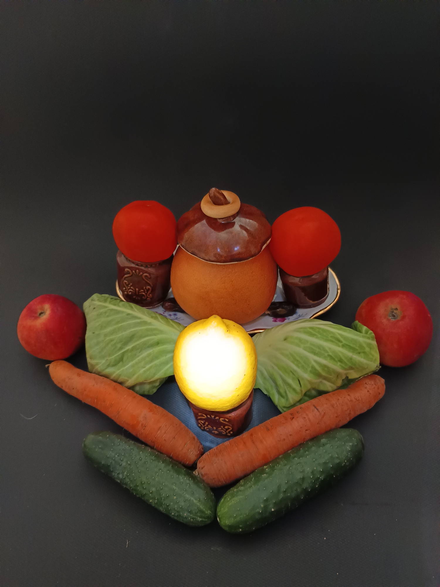 The image shows a still life arrangement of fruits and vegetables on a dark, textured surface, likely a table. The arrangement is symmetrical and appears to be set against a dark, plain background.
In the center, a rustic, orange-brown ceramic pot with a lid is positioned. To its left and right, on small, ornate saucers, are two small dark brown jars topped with red tomatoes. Behind these, and slightly obscured by the central pot, are two wedges of green cabbage, fanning out.
In the foreground, a bright yellow lemon, cut in half and illuminated from within, is placed centrally, resting on a small, patterned dark brown ceramic base. Flanking the lemon, and extending outwards in a fan shape, are two large, orange carrots. Beneath the carrots, two green cucumbers are positioned. On the outer edges of the arrangement, two red apples are visible, one on the left and one on the right.
The lighting is somewhat dramatic, with a strong light source illuminating the lemon from within and casting shadows. The overall impression is one of a stylized, rustic presentation of fresh produce.
There is no information or visual evidence to suggest the location of this scene in the city of Tyshky, Ukraine. The image solely depicts the arrangement of objects.