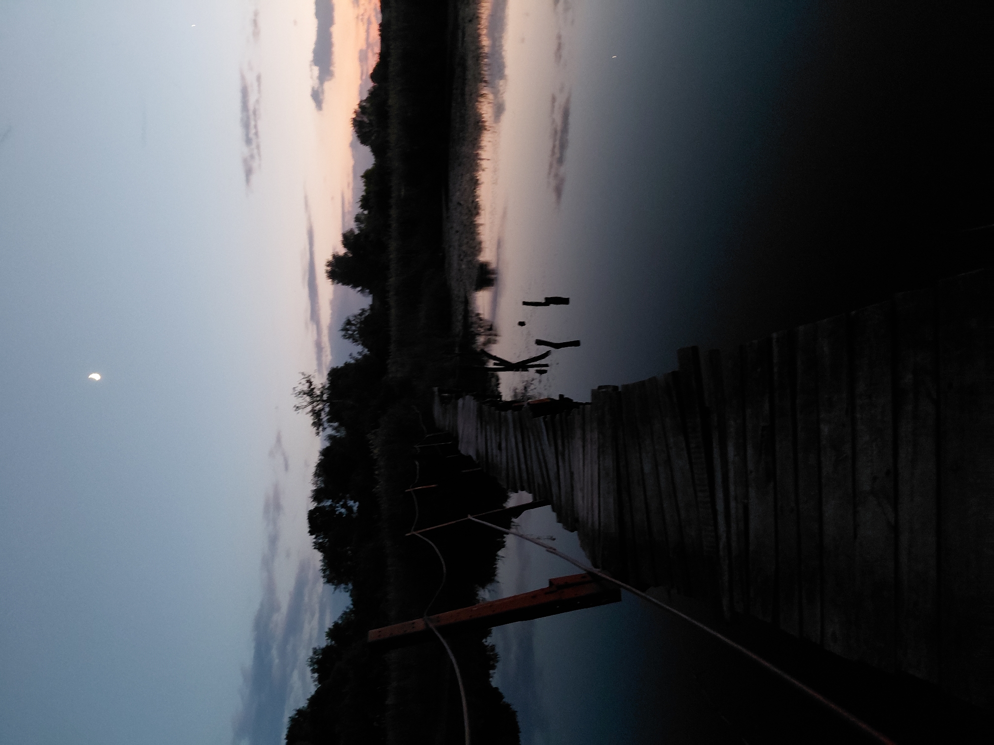 This image depicts a wooden pier extending over dark water at dusk. The sky is a gradient of deep blue at the top, transitioning to lighter blues and pinks near the horizon, with scattered clouds. A crescent moon is visible in the upper sky. The pier, made of weathered wooden planks, is in the foreground and leads towards a dark treeline in the background. The water reflects the sky and surrounding vegetation. The scene appears to be a natural environment, possibly a lake or river, near the city of Ustyvytsia, Ukraine. There are no people visible in the image. The overall atmosphere is serene and twilight.