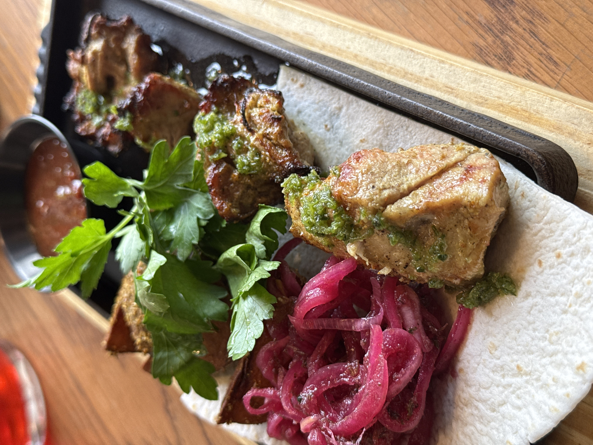The image displays a close-up of a rustic food presentation, likely a meal in a restaurant or eatery. The main subjects are several pieces of grilled or pan-fried meat, possibly pork or chicken shashlik, seasoned with a vibrant green herb sauce or marinade. These meat pieces are arranged on a piece of white flatbread or lavash, which sits on a dark, heavy-looking serving tray. The tray, possibly cast iron, is placed on a light wooden board.
Accompanying the meat are bright pink, thinly sliced pickled red onions, and a generous sprig of fresh green parsley or cilantro, adding color and freshness to the dish. To the left, a small metal dish contains a reddish-brown dipping sauce.
The overall scene suggests a casual dining experience. The food appears freshly prepared and appetizing. The lighting is indirect, typical of an indoor setting, and does not indicate a specific time of day. No people are visible in the frame, and there is no discernible text. The presentation style, with the grilled meat, pickled onions, and flatbread, aligns with popular dishes found in the Votkinsk, Russia region. A blurred red object, possibly a drink in a glass, is visible in the bottom left corner.