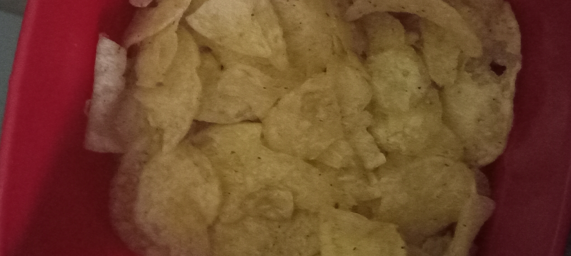 The image shows a close-up view of potato chips in a red container. The chips are golden brown and appear to be slightly greasy. They are piled on top of each other, filling the container. The background is out of focus, but appears to be a dark surface. There are no people or other objects visible in the image. The image does not contain any information about the location in the city of Koviahy, Ukraine.