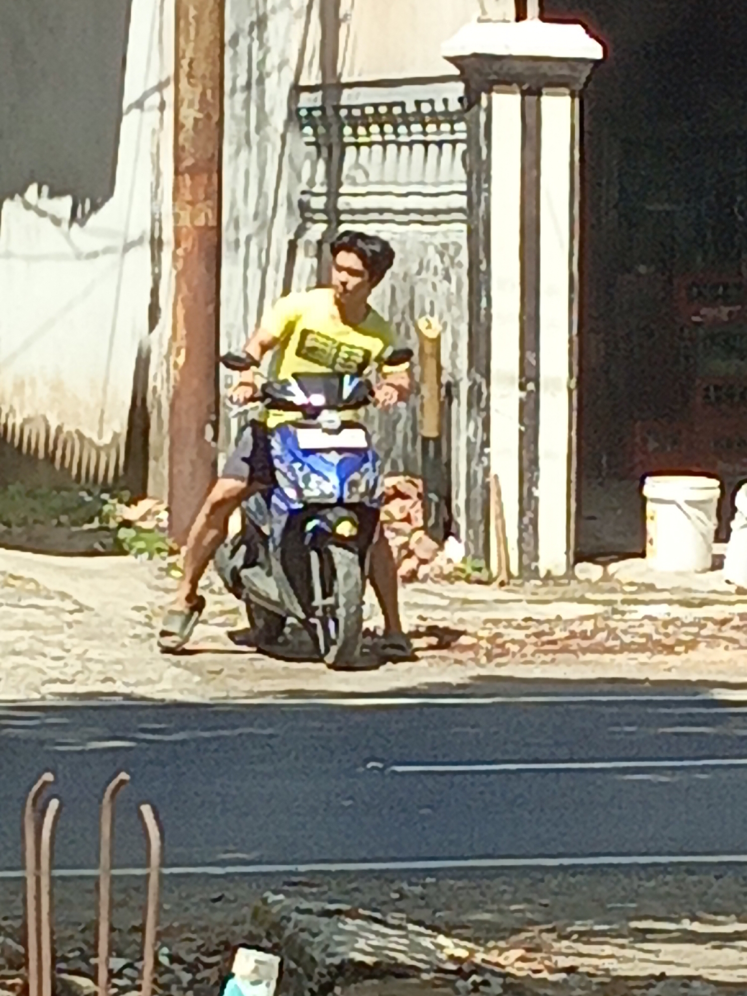 A young man in a bright yellow t-shirt is sitting on a motorcycle with blue and white accents. His legs are positioned on either side of the motorcycle. The man is looking forward and appears to be ready to ride. The motorcycle is parked on a dusty surface in front of a building with a textured, light-colored wall and a decorative pillar. In the background, there are some white buckets. The scene appears to be outdoors, possibly on a street in Srono, Indonesia, given the presence of a road in the foreground.