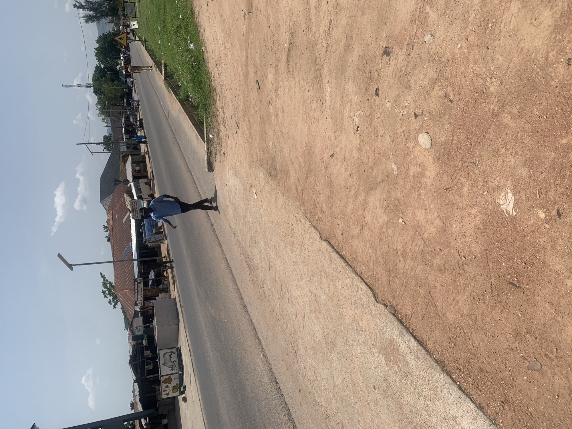 The image depicts a street scene in Jalingo, Nigeria, with a person walking on the side of the road. The road itself is paved and appears to be a main thoroughfare, with buildings and market stalls lining one side. The other side of the road is a dirt embankment covered in sparse grass and debris.
The person, wearing a blue shirt and dark pants, is walking away from the camera, towards the left of the frame. They are the primary subject in the foreground. In the background, there are various market stalls, some with corrugated metal roofs. A street lamp is visible, suggesting it is daytime.
The sky is blue with scattered clouds, indicating fair weather. The overall impression is of a bustling street in a developing urban area. The time of day appears to be late morning or early afternoon due to the lighting. There is no discernible text visible in the image. The scene is likely a typical day in this Nigerian town, with commercial activity and people going about their daily routines.