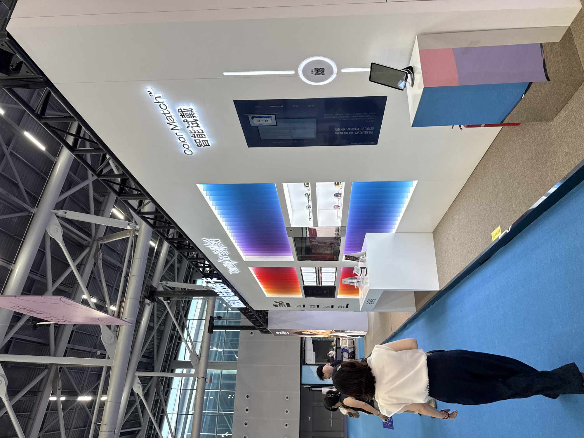 The image depicts an indoor exhibition booth at a trade show. The booth has a modern, white wall structure adorned with a prominent neon sign that reads "Color Match" in English and Chinese characters below. The wall also features a large digital display showing a user interface, a QR code for scanning, and a tablet device mounted on a cube-shaped stand. Several shelves behind the wall display what appear to be eyeglass frames, illuminated by a gradient of colored lights ranging from red to purple and blue.
In the foreground, a woman wearing a white sleeveless top and dark pants is walking away from the camera on a vibrant blue carpeted floor. Other attendees are visible in the background, including a man in a black shirt and another woman with dark hair. The overall environment suggests a commercial exhibition or trade fair, likely in a large convention center. The presence of a fire extinguisher near the display further points to safety measures common in such venues. The context strongly suggests the event is taking place in Haizhu District, China, as indicated by the prompt.