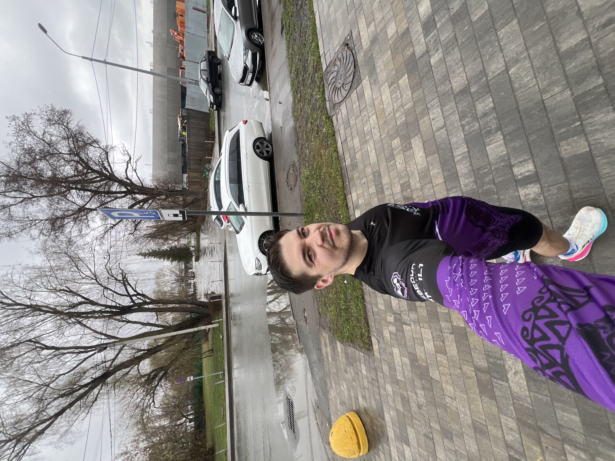 A man is standing on a paved sidewalk on a cloudy day. He is wearing a black shirt with purple patterned sleeves and leggings, and white running shoes. The sidewalk is made of gray rectangular blocks. A yellow bollard is visible near the man's feet. To the left of the man, there is a wet street with several parked cars, including a white sedan. Behind the cars, there is a grassy area and bare trees. In the background, there is a tall, gray building. A street light pole is also visible. The man is looking at the camera and smiling. Based on the style of buildings and street furniture, the location is likely in an urban area, possibly Kazan, Russia.